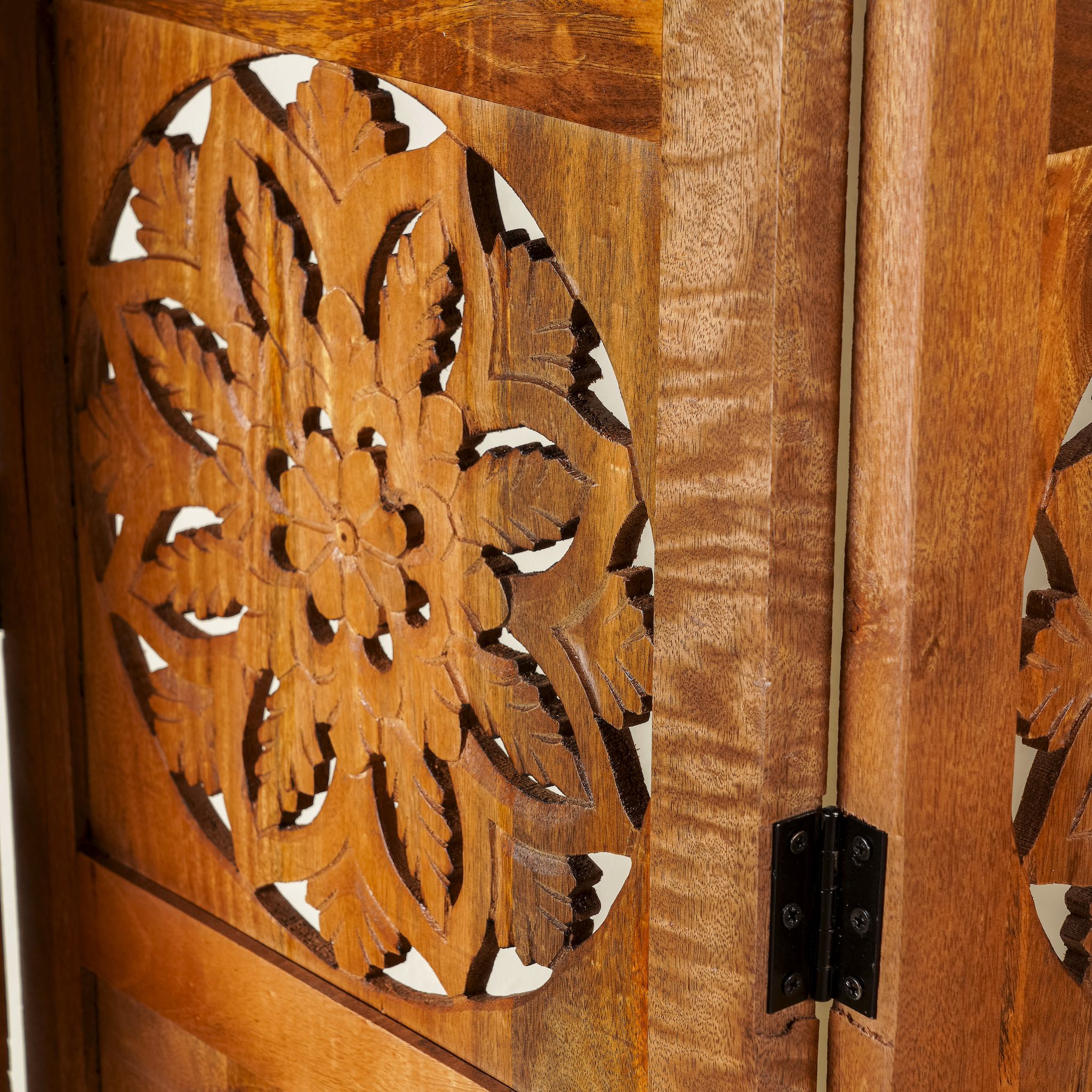 Side angle image of a foldable room partition made of mango wood, featuring four panels with symmetrical hand-carved circular floral medallions, finished in a rich natural wood tone and connected by black metal hinges for easy folding and adjustment.