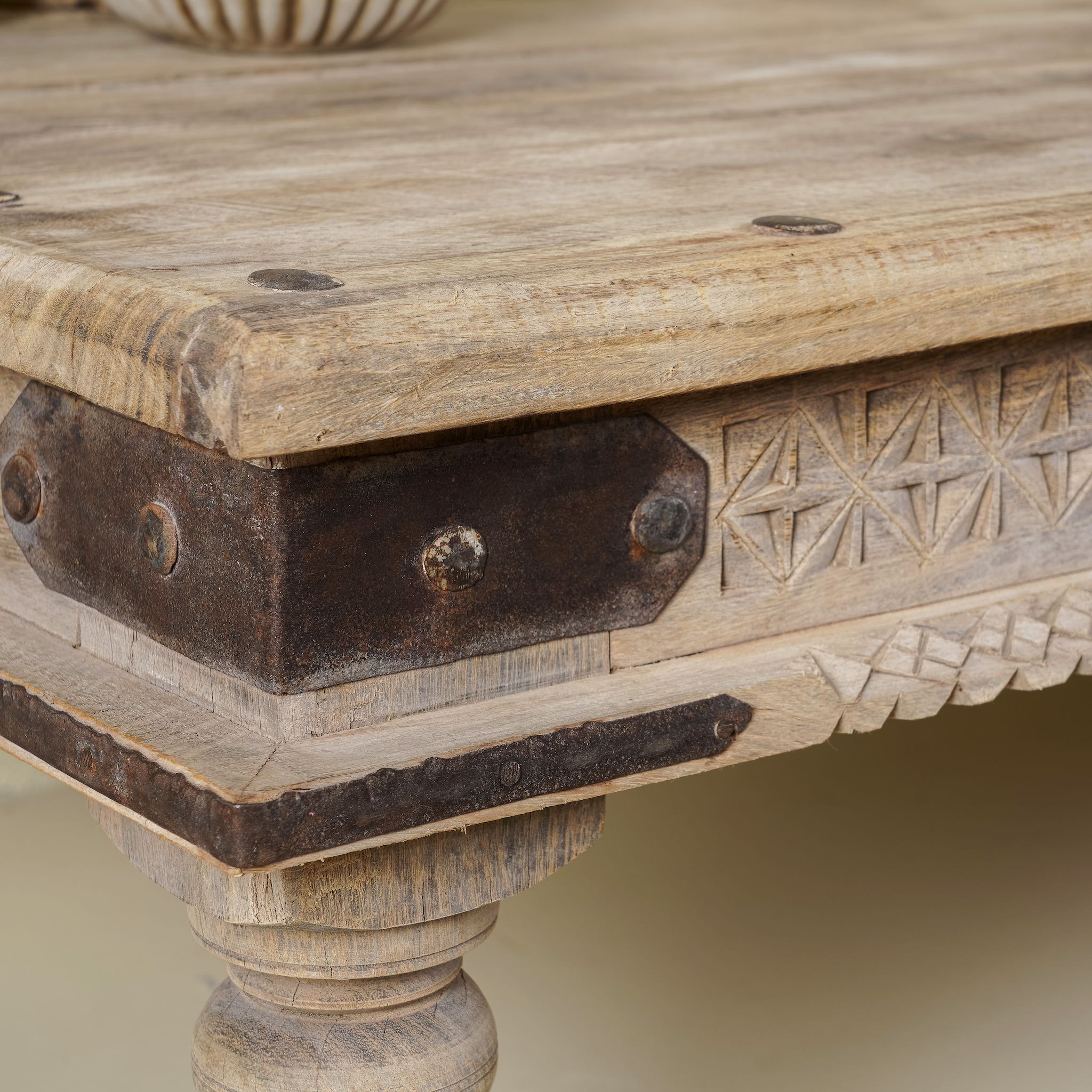 Close up of a corner of the rustic coffee table Amana made from old reclaimed teak wood, featuring a weathered finish, turned legs, decorative carved apron, and visible metal rivets, styled with earthy decor in a minimal interior setting.