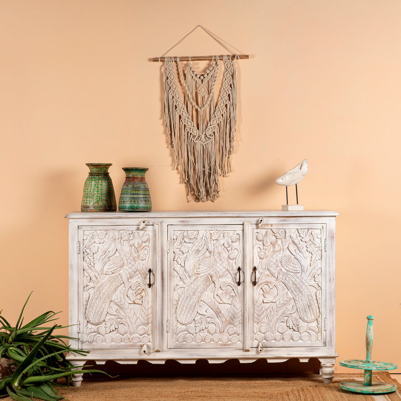 A hand-carved mango wood sideboard with a whitewashed finish, featuring intricate peacock motifs. The sideboard has three doors and is placed against a wall with a decorative hanging above it.