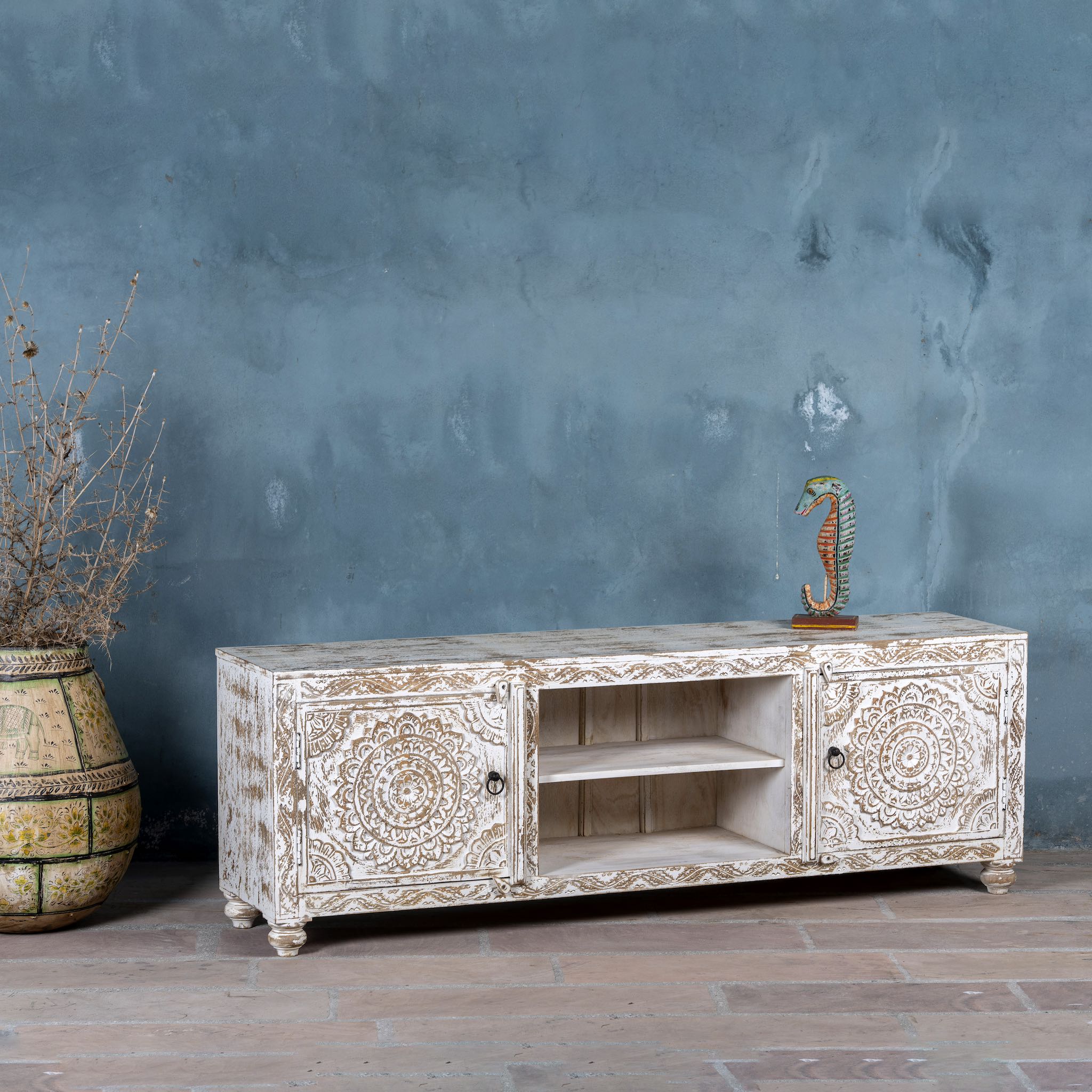 Side angle image of a white washed mango wood lowboard with carved floral doors and open shelving, 180 × 40 × 60 cm.