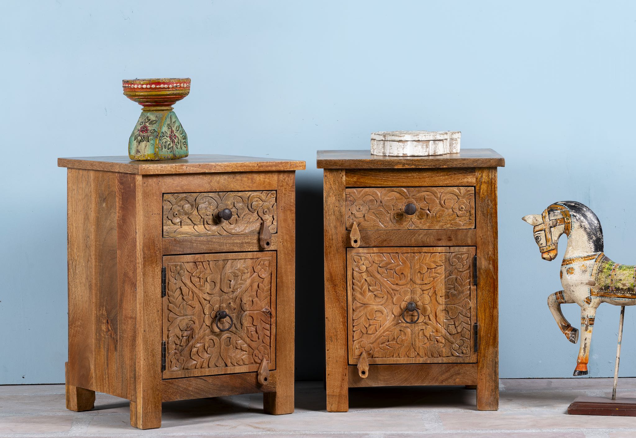Side angle image of a pair of mango wood night stands with floral hand carvings in natural wood colour.