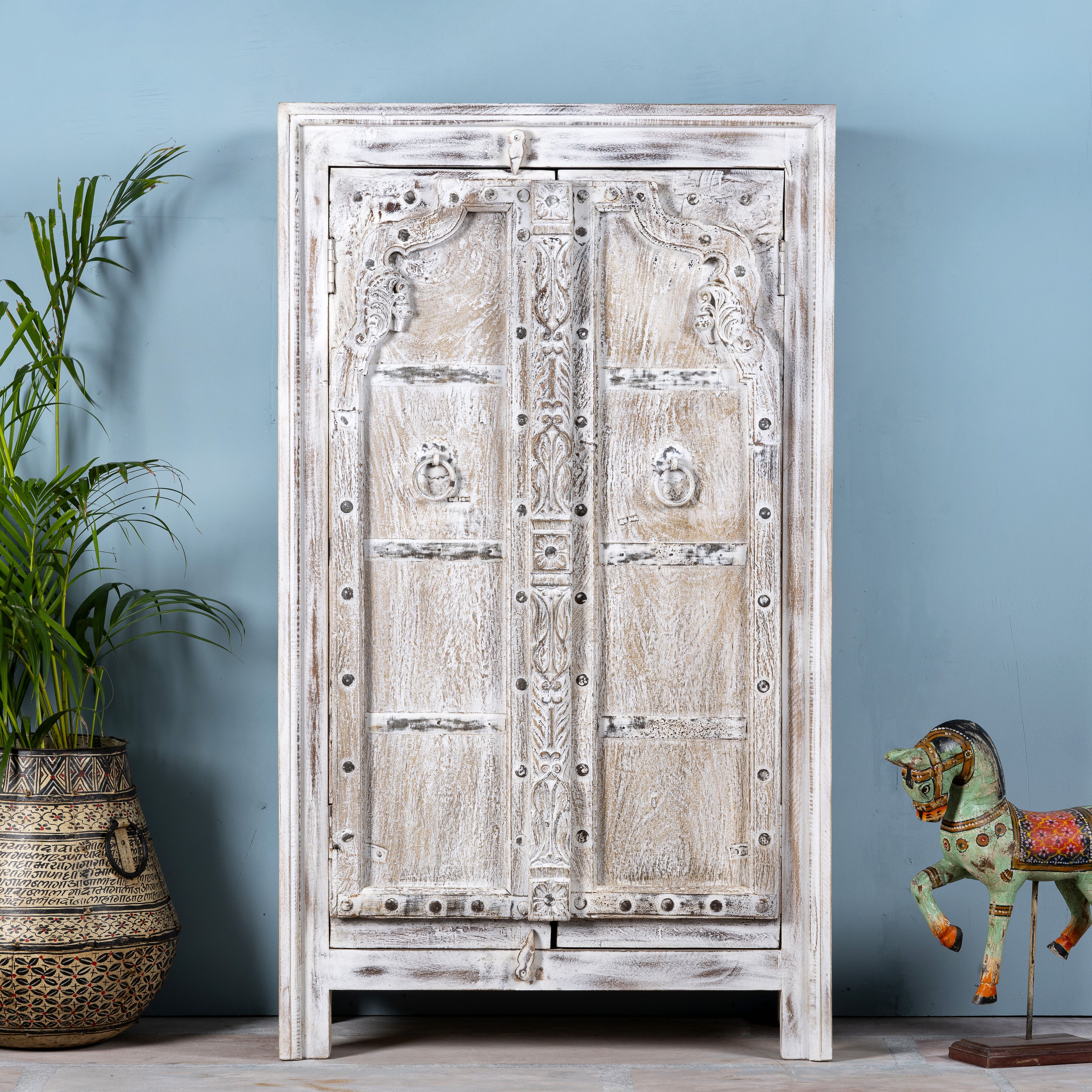 A white-washed, wooden cabinet with intricate iron applications, standing in a room with a potted plant and a decorative horse figure.