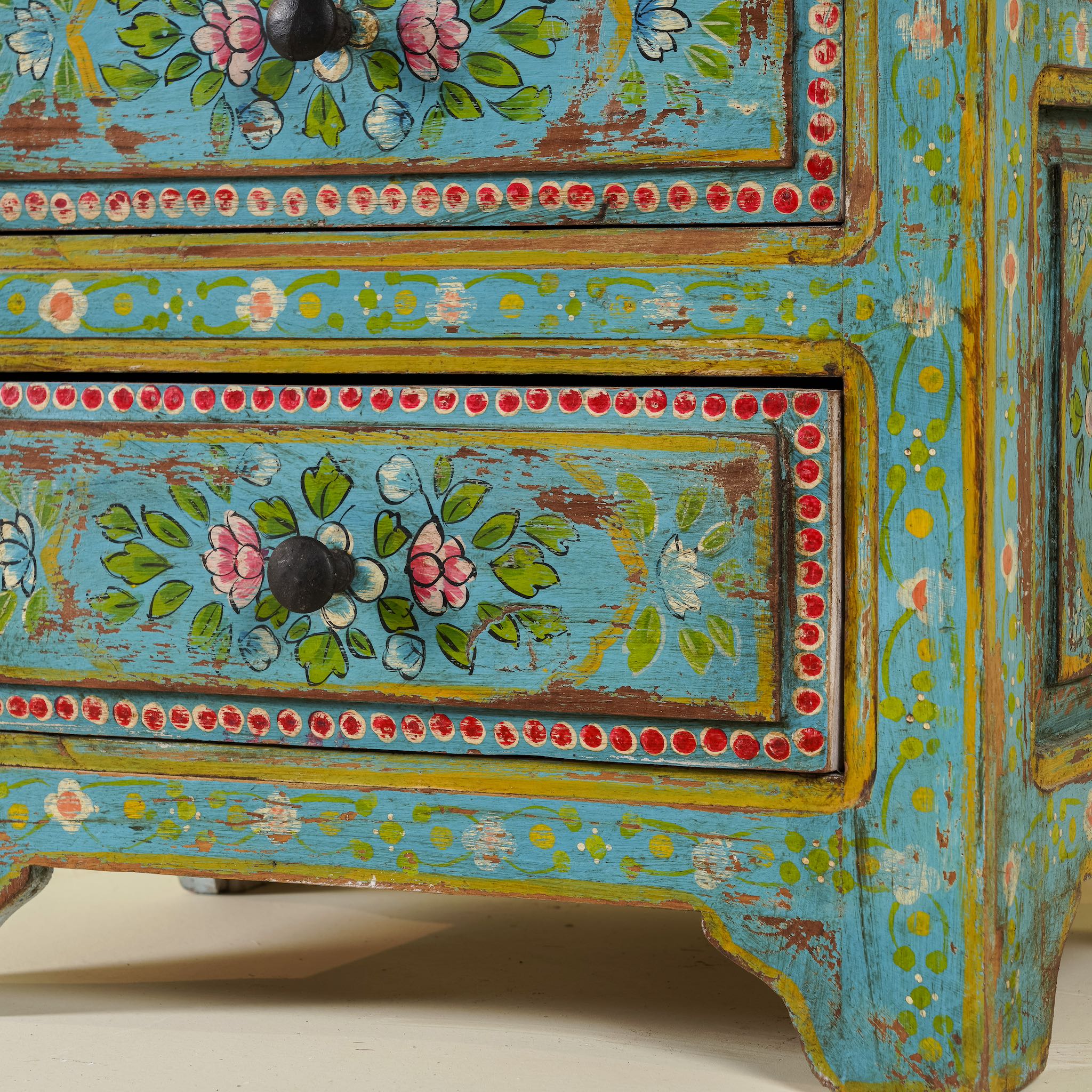 Close up image of the leg part of a hand-painted reclaimed wood nightstand with vibrant colours and floral patterns, featuring three drawers, placed next to a vase with dried plants.