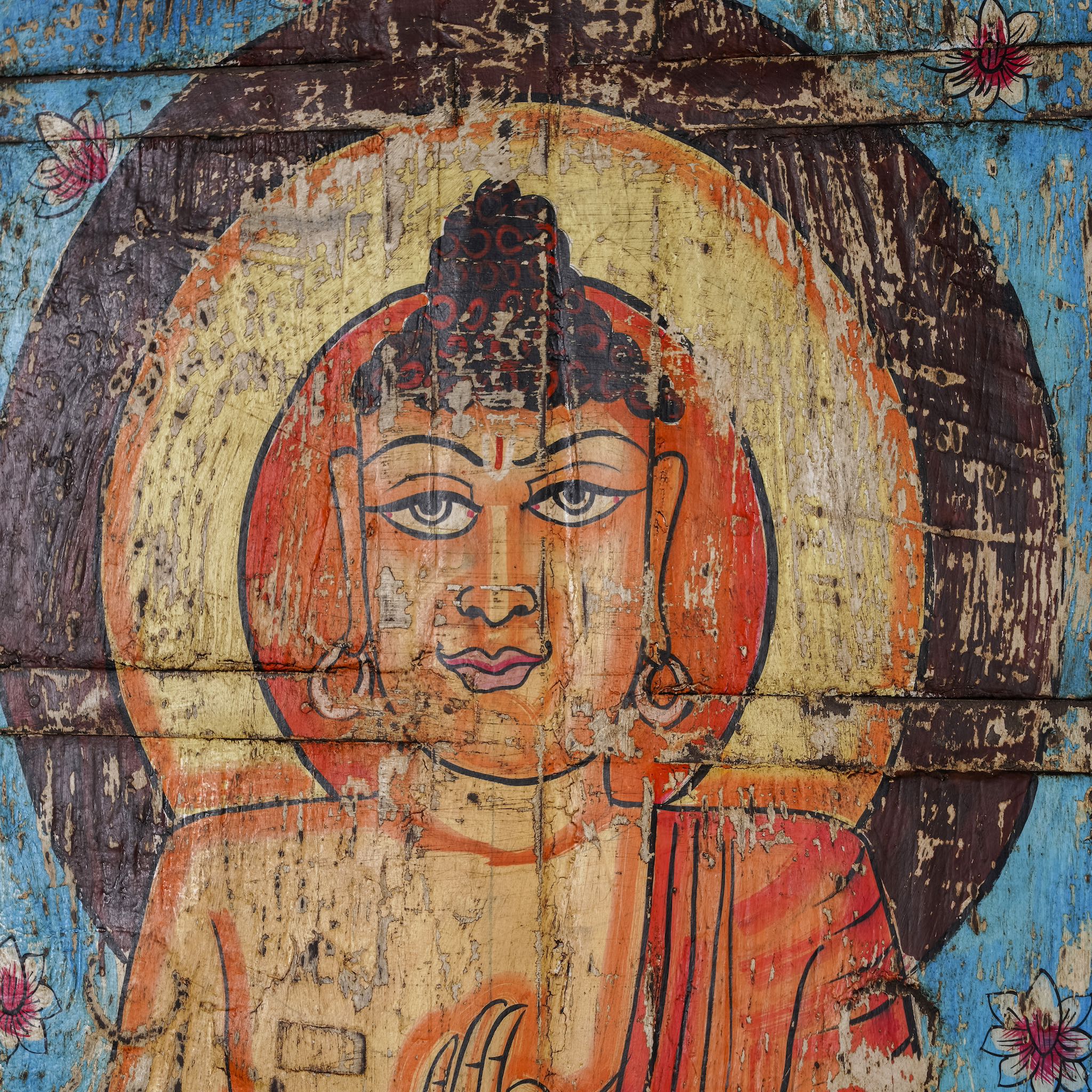 Close up image of a hand-painted wall panel made from reclaimed wood, featuring a vibrant depiction of Buddha in a meditative pose on a lotus flower, with a bright blue floral background and a worn, rustic finish that adds vintage charm.