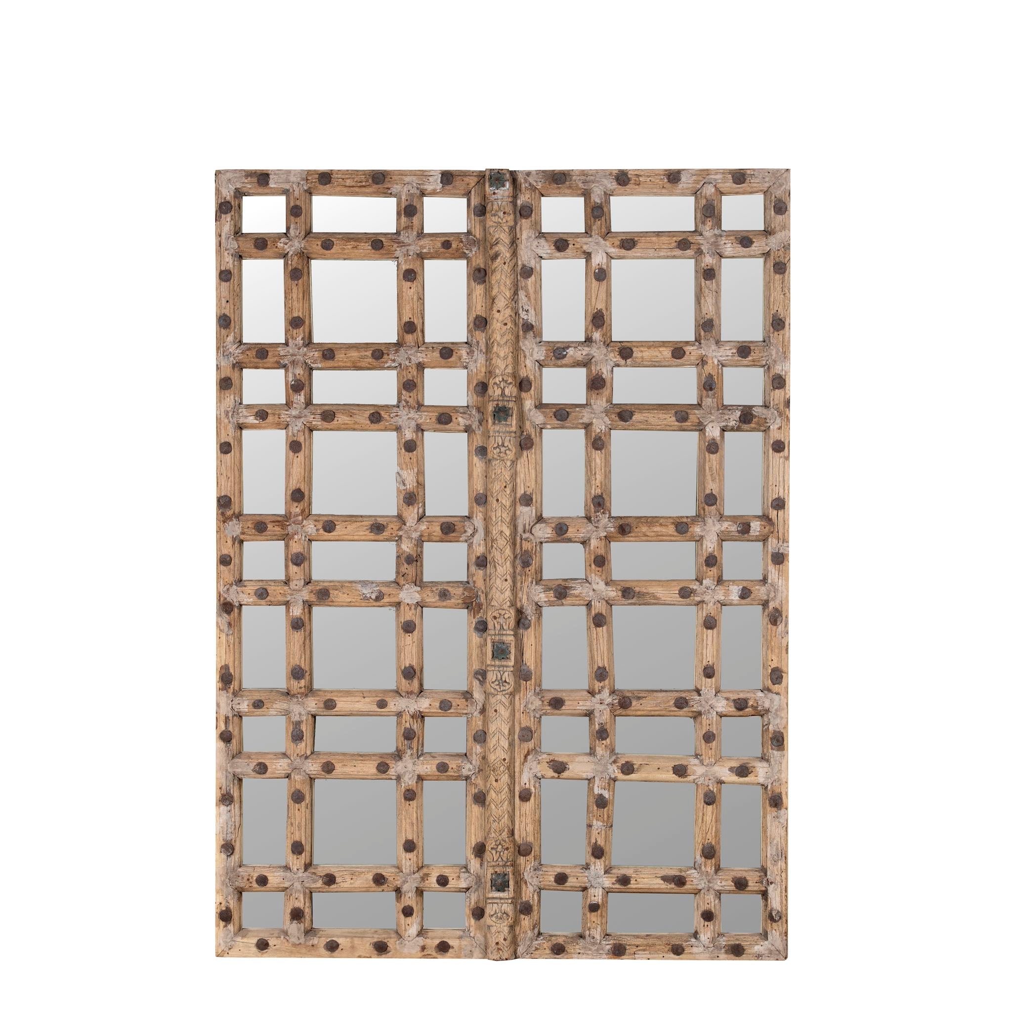 White background picture of a statement wall mirror made from an old teak window frame with large square grid pattern, metal studs, and decorative center carving — a bold blend of rustic charm and historic craftsmanship.