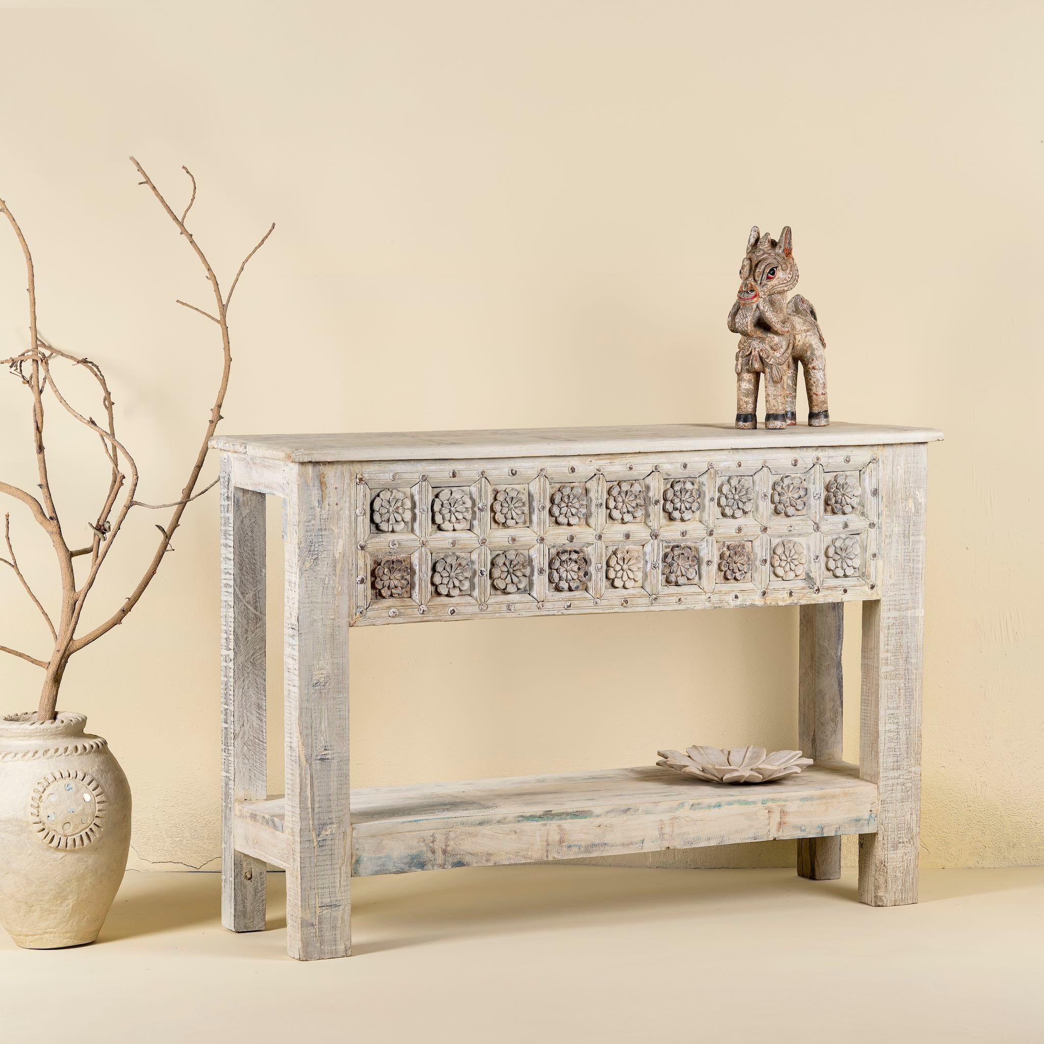 Side angle of a handcarved console table made from reclaimed teak wood, featuring a detailed front panel with geometric floral motifs and a distressed whitewashed finish, complemented by a lower shelf for added storage or display.