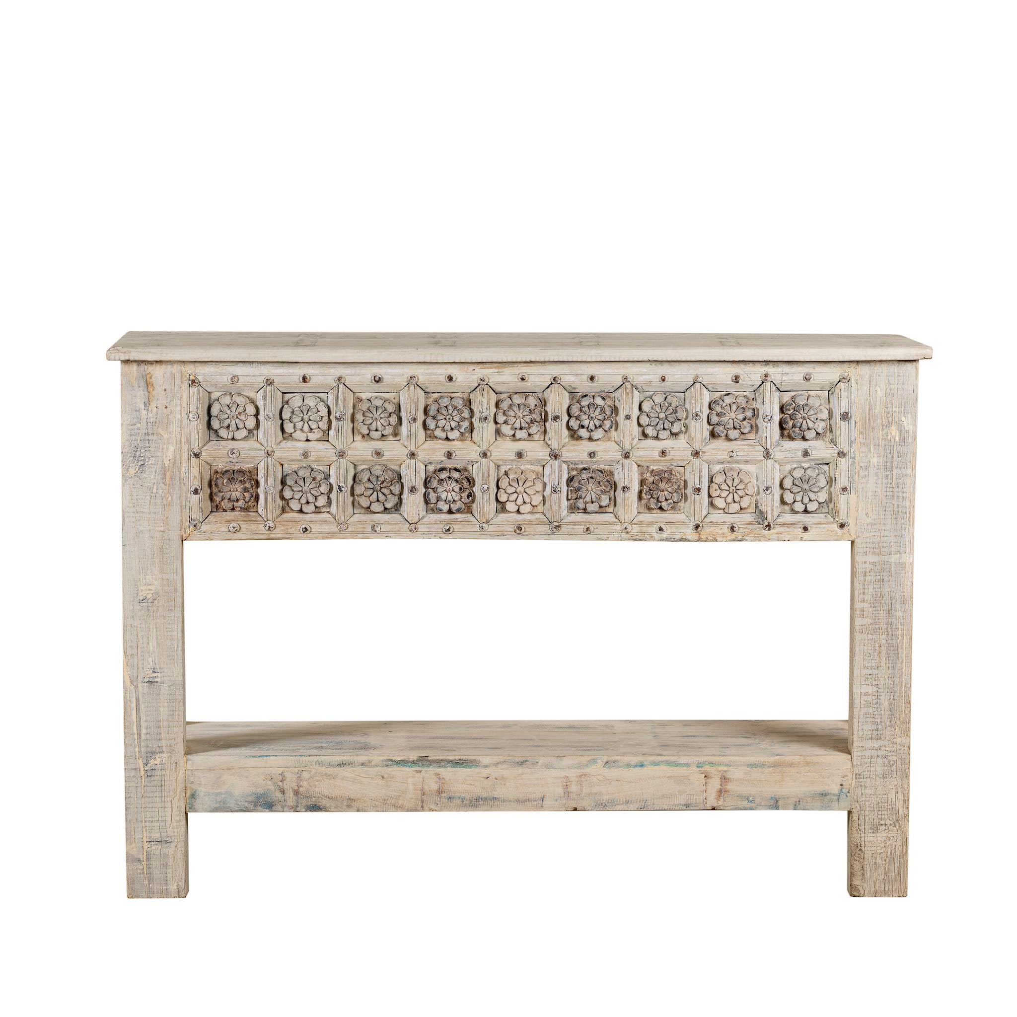 White background image of a hand carved console table made from reclaimed teak wood, featuring a detailed front panel with geometric floral motifs and a distressed whitewashed finish, complemented by a lower shelf for added storage or display.