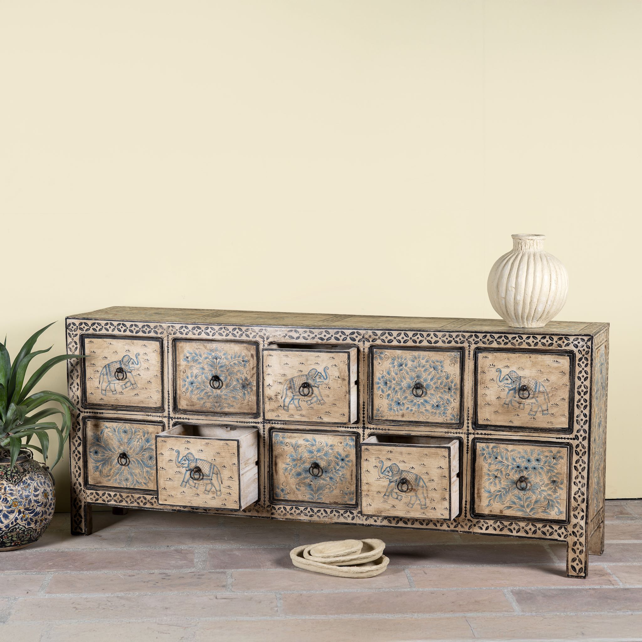 Side angle image with open drawers of a hand painted mango wood chest of 10 drawers with elephant and floral motifs; hand painted furniture for a boho chic living room, 200 × 41 × 85 cm.