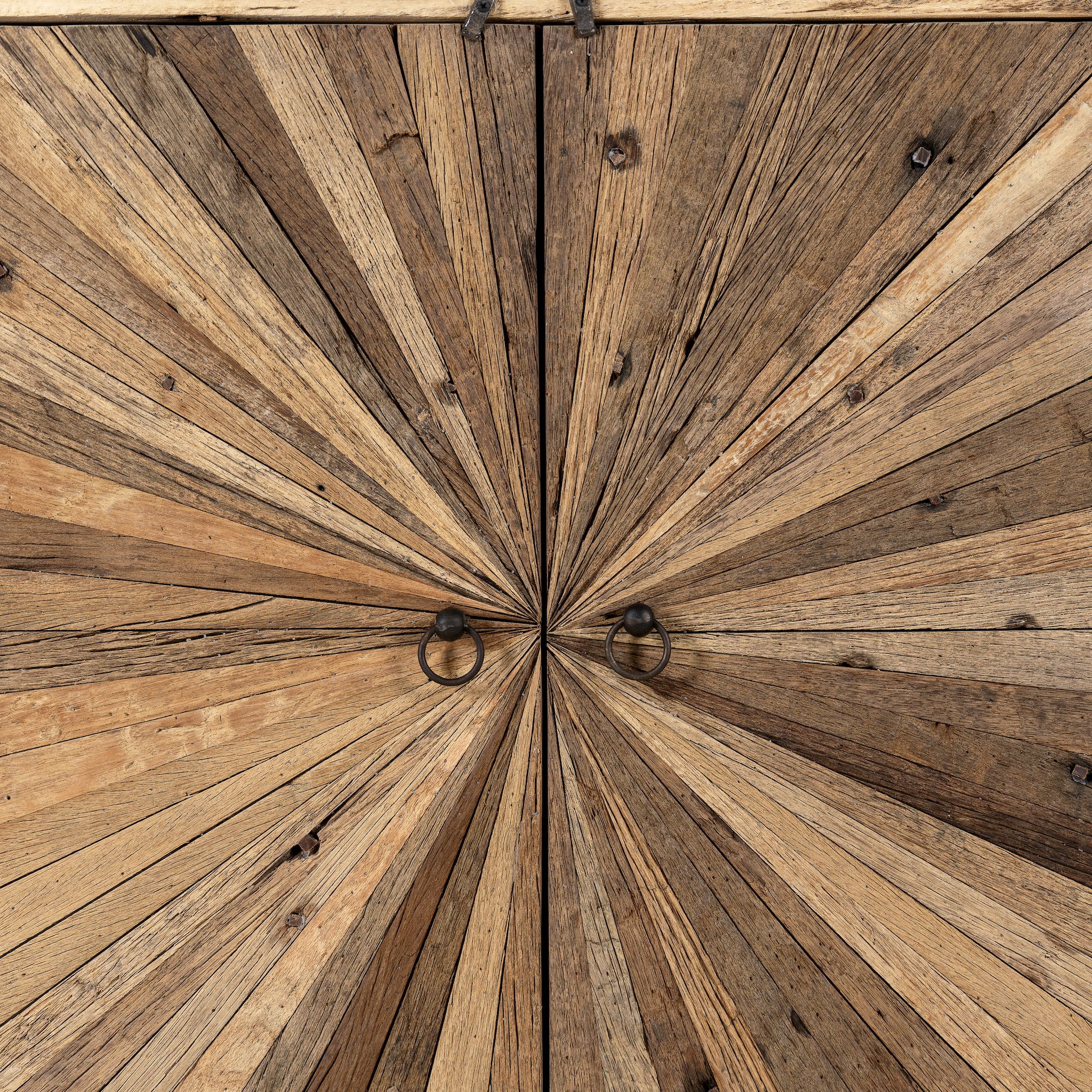 Close up image of a artistic wooden cabinet in rustic style, made from reclaimed wood with a striking sunburst pattern on the double doors, featuring subtle color variations and round metal ring pulls, elevated on rounded wooden feet.
