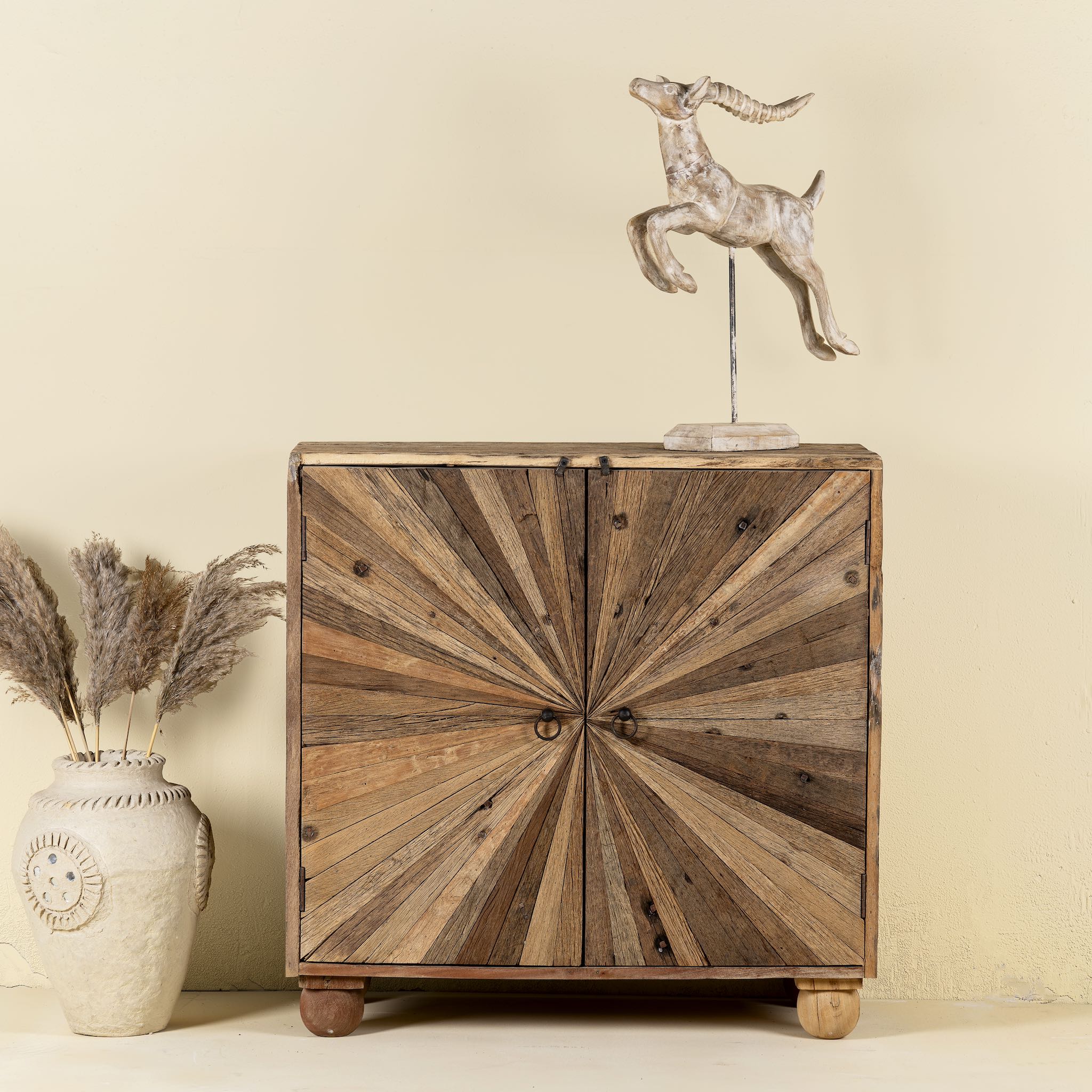 Front view image of a artistic wooden cabinet in rustic style, made from reclaimed wood with a striking sunburst pattern on the double doors, featuring subtle color variations and round metal ring pulls, elevated on rounded wooden feet.