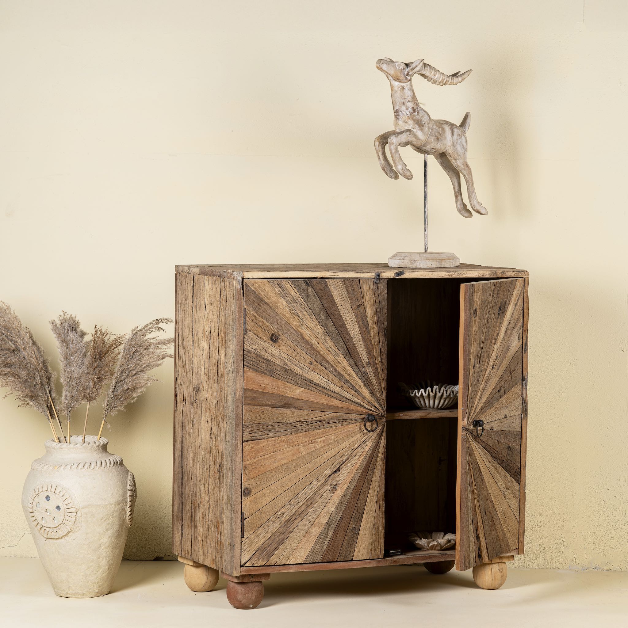 Open door image of a artistic wooden cabinet in rustic style, made from reclaimed wood with a striking sunburst pattern on the double doors, featuring subtle color variations and round metal ring pulls, elevated on rounded wooden feet.