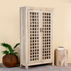 Side angle image of a sandblasted mango wood showcase with two glass-panelled grid doors and four interior compartments, finished in a light sun-washed tone, ideal as living room furniture.