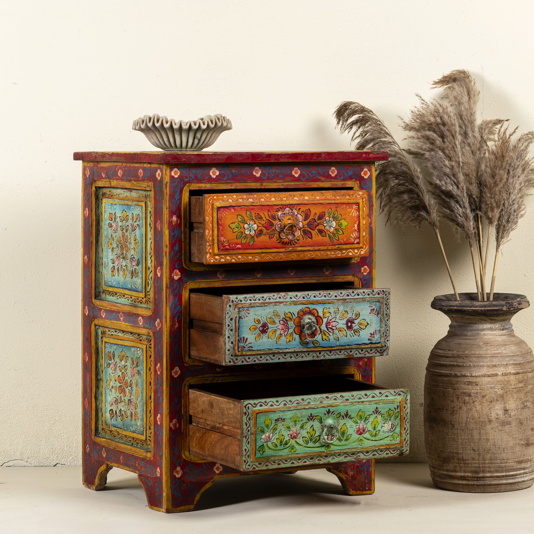 Side angle image with open drawers of a hand-painted reclaimed wood nightstand with multiple drawers, featuring vibrant colours and floral patterns, placed next to a vase with dried plants.