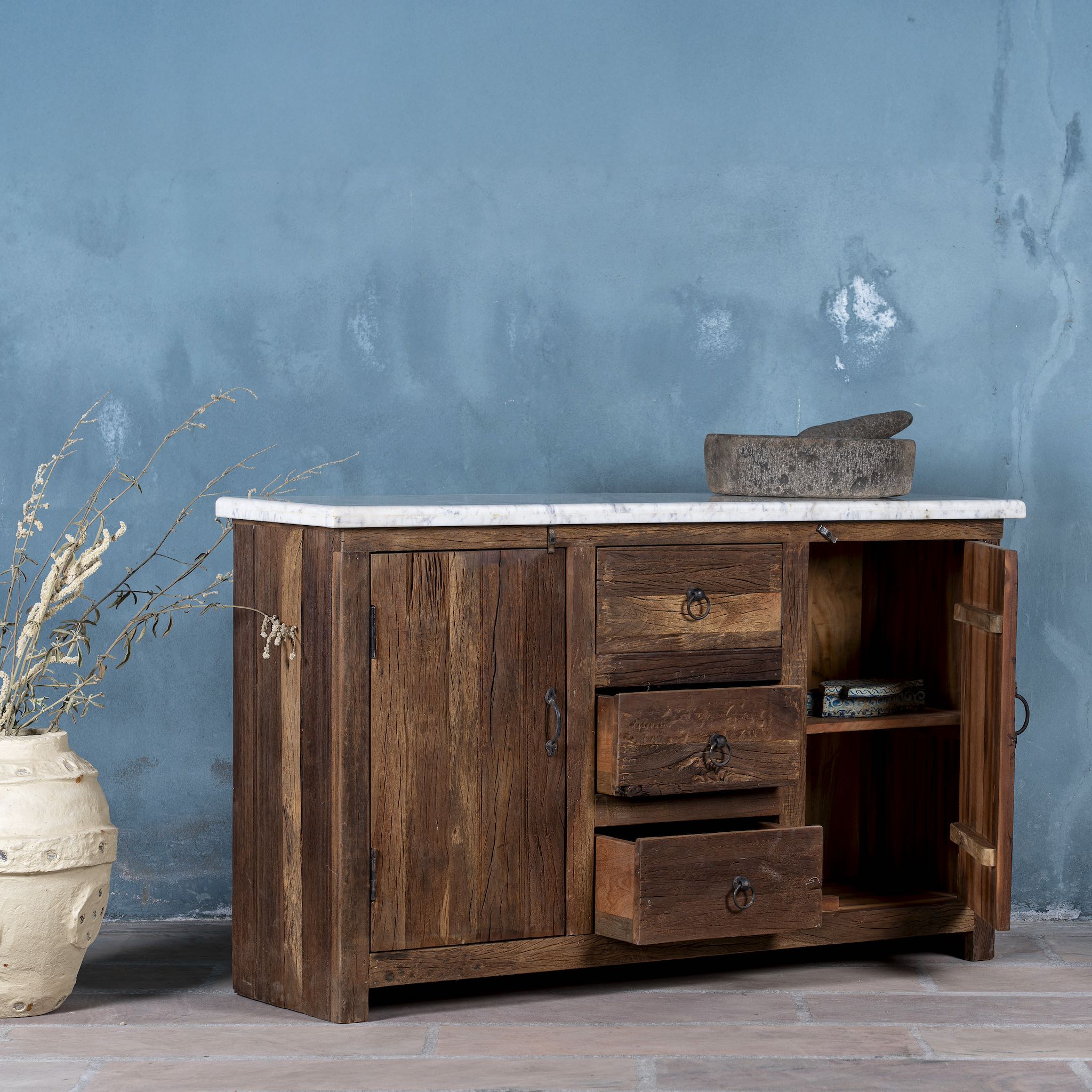 Open door view of a driftwood sideboard with iron handles, 3 drawers and 2 doors and a slidable marble top in a maritime scene.