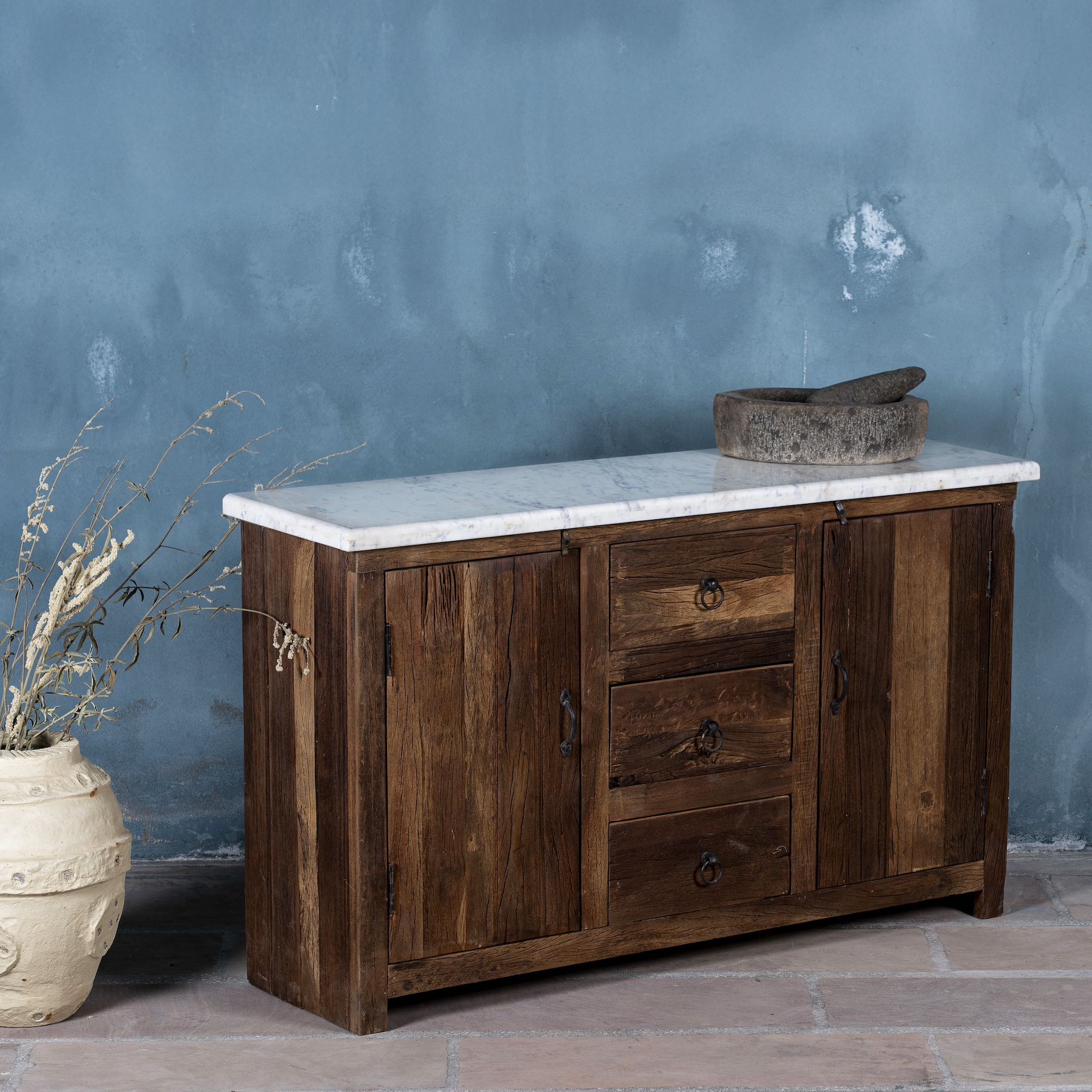 Side view of a driftwood sideboard with iron handles, 3 drawers and 2 doors and a slidable marble top in a maritime scene.