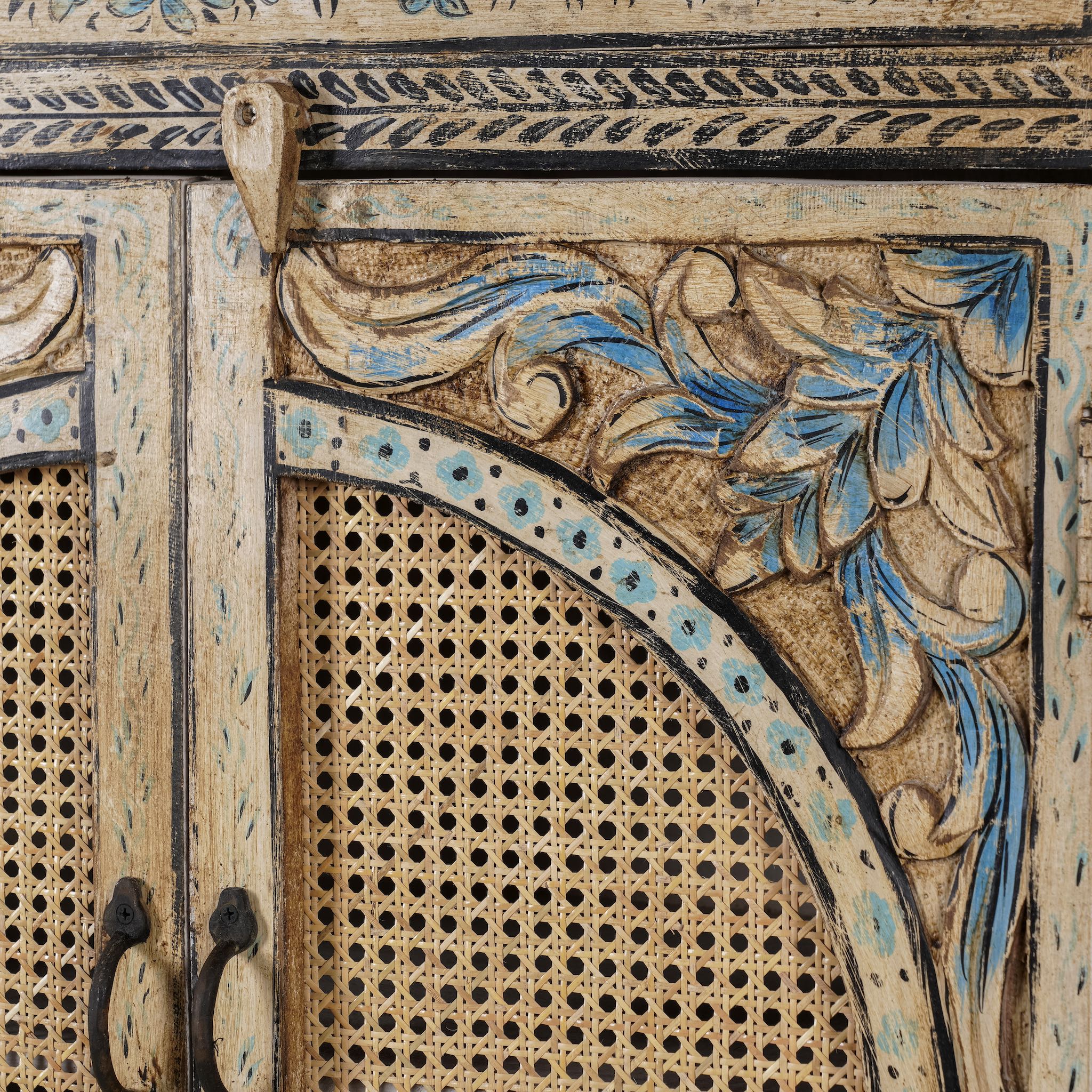 Close up image of a hand-painted cabinet made from mango wood, featuring rattan inlay doors framed by a carved circular motif, floral blue and green accents, and five drawers for additional storage.