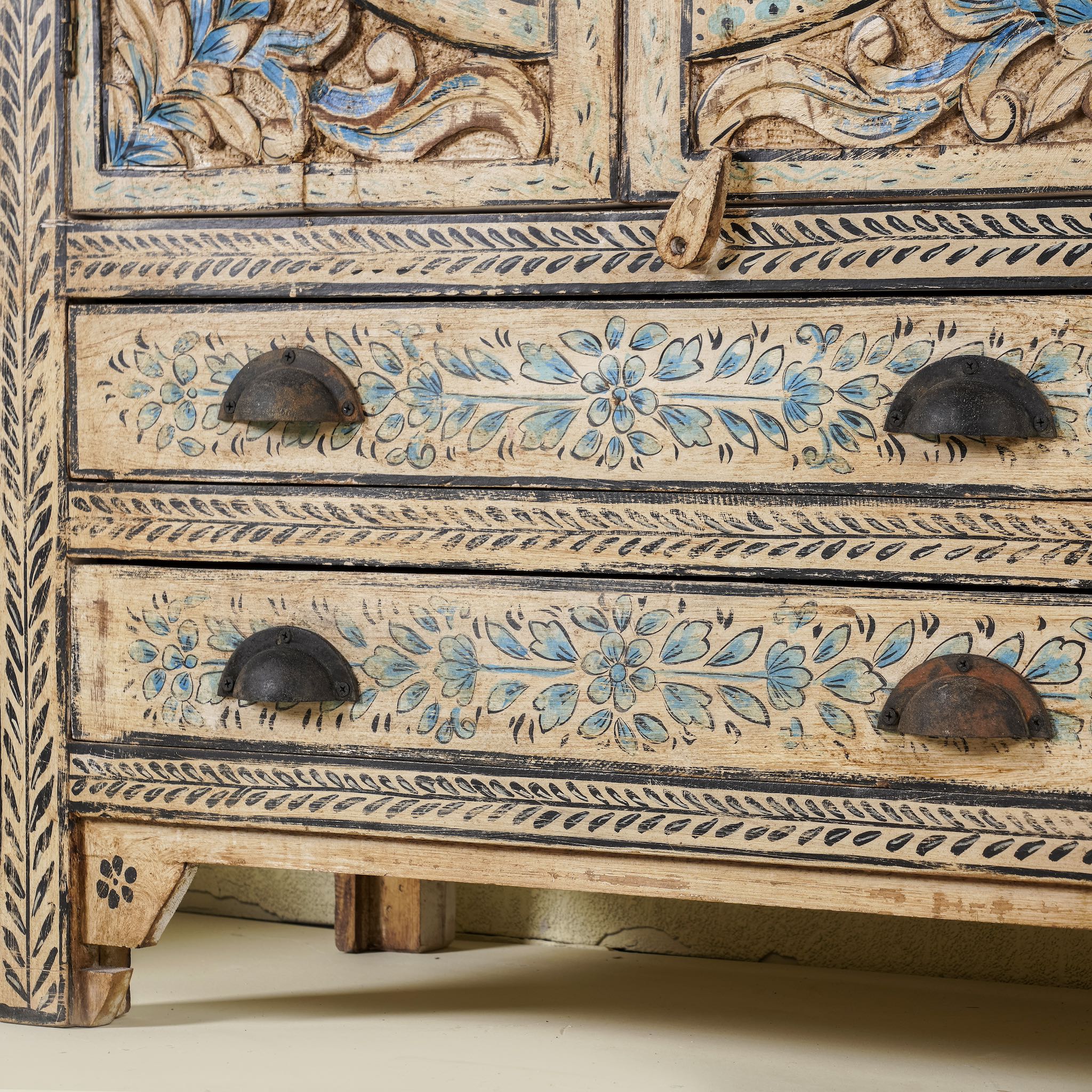 Close up image of the leg part of a hand-painted cabinet made from mango wood, featuring rattan inlay doors framed by a carved circular motif, floral blue and green accents, and five drawers for additional storage.