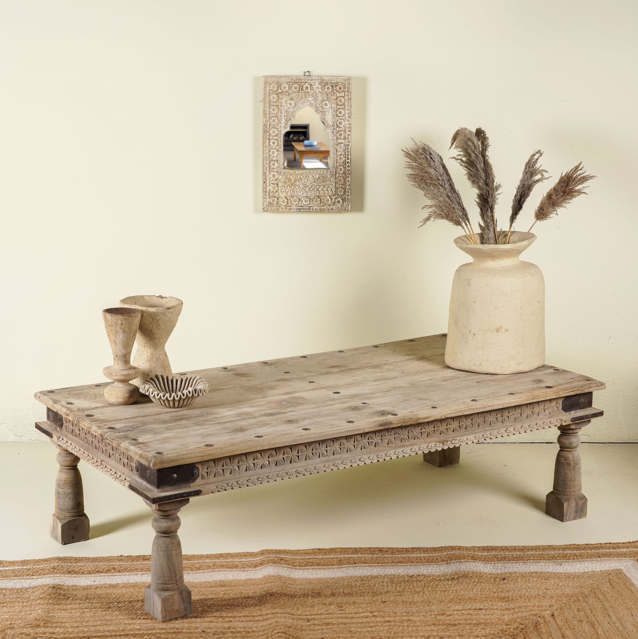 Side angle of a rustic coffee table made from old reclaimed teak wood, featuring a weathered finish, turned legs, decorative carved apron, and visible metal rivets, styled with earthy decor in a minimal interior setting.