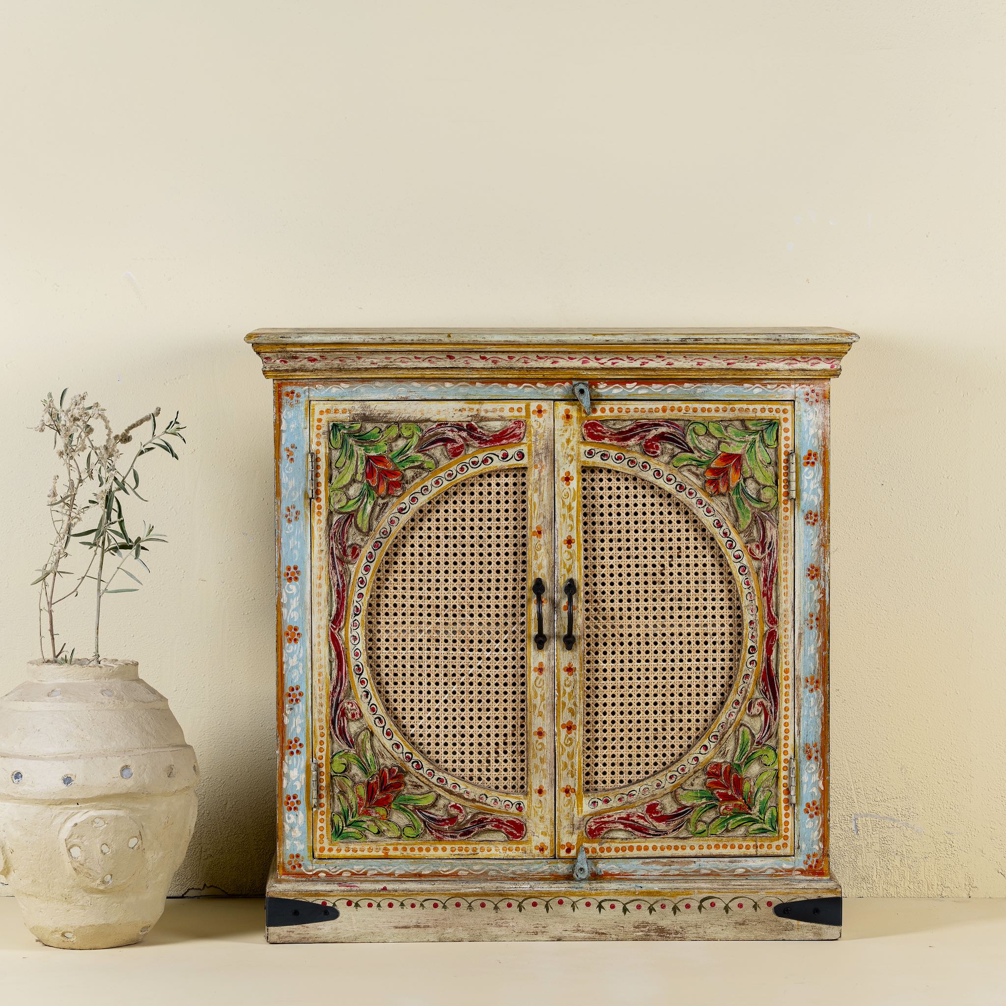 Front view of a hand-painted, hand-carved cupboard made of mango wood with floral design and rattan inserts.