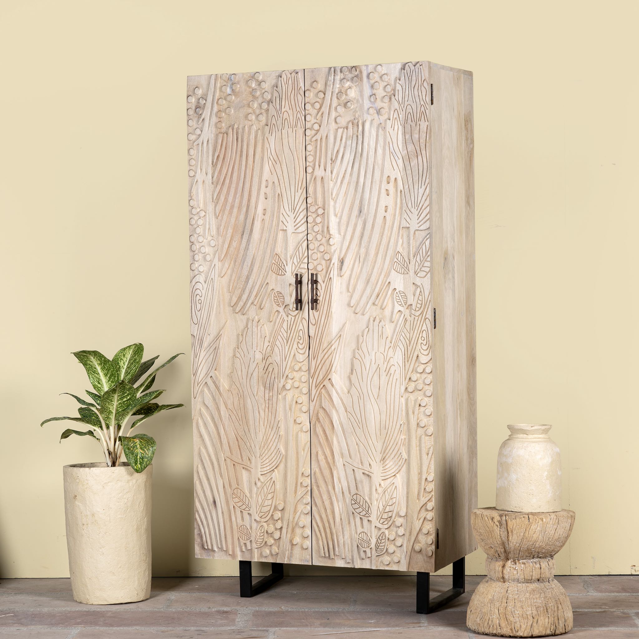 Side angle image of a hand-carved mango wood wardrobe with sandblasted finish and iron stand; artisanal bedroom furniture, 90 × 46 × 183 cm.