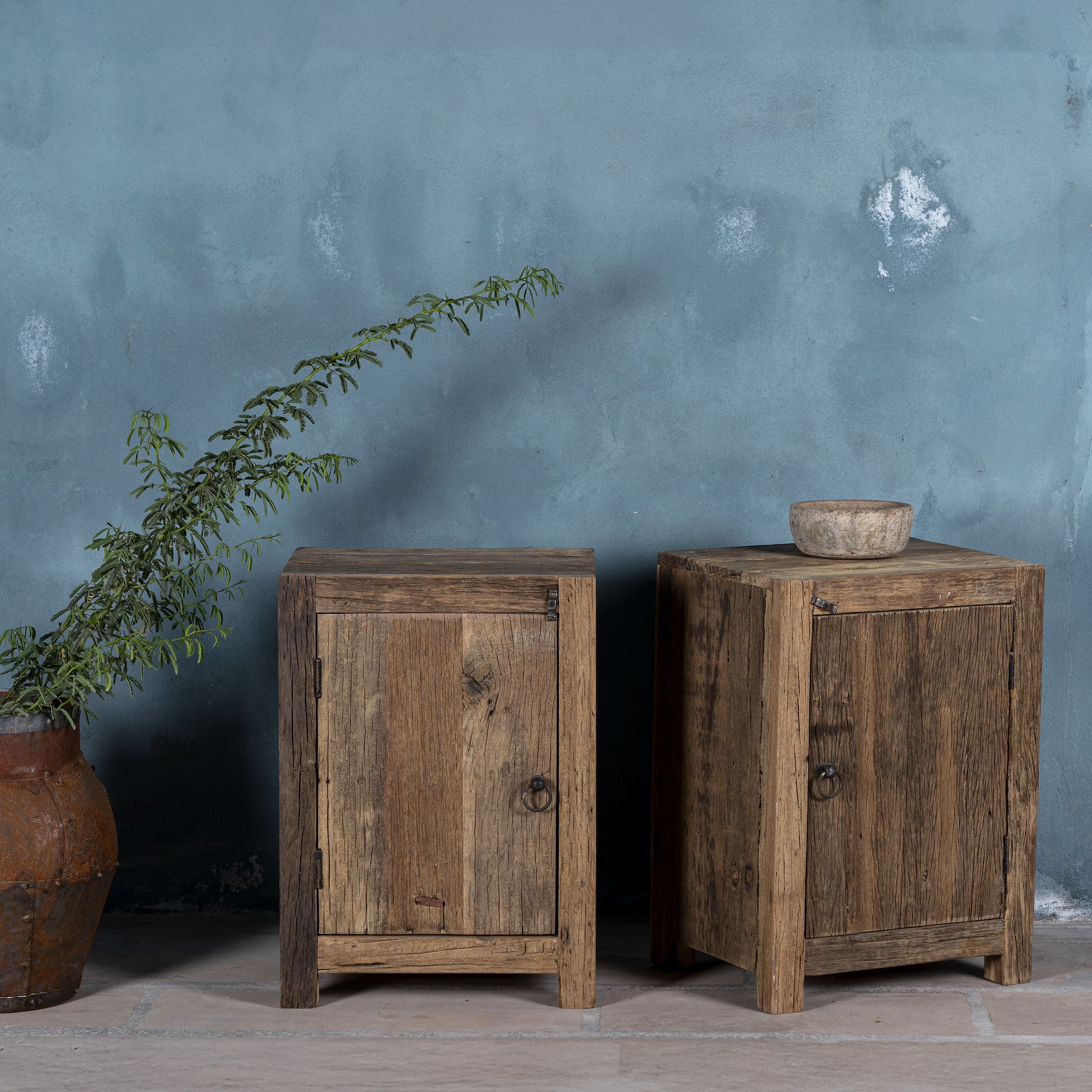 Side view of a pair of night stands in rustic minimalistic design with iron door handles decorated in front of a blue wall.