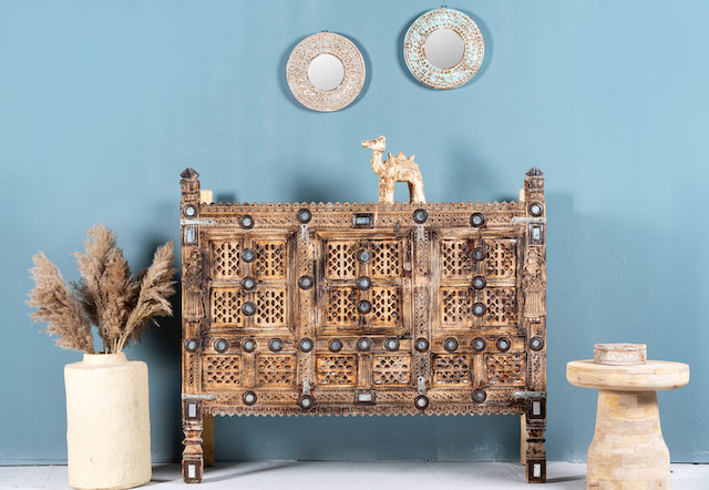 An antique Indian treasure chest with sandblasted surface and iron applications, featuring intricate hand-carved patterns, displayed against a blue wall with decorative items around it.