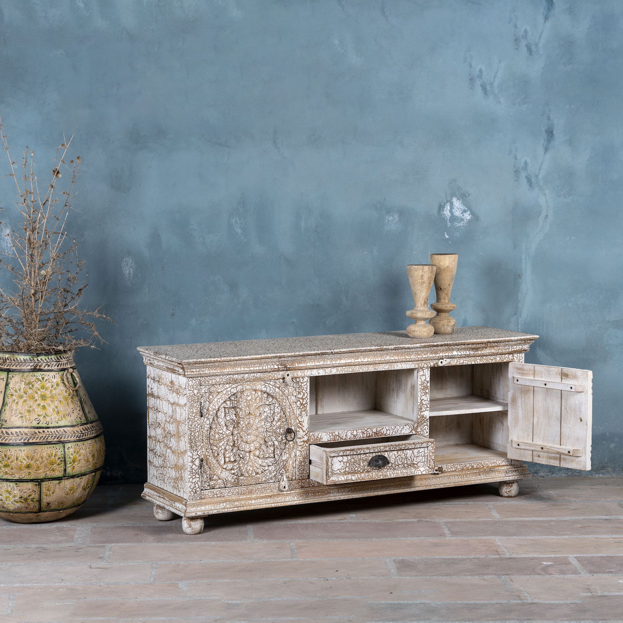Side angle view of a carved mango wood lowboard with floral doors, crackled vintage finish, drawer, and open shelf, 150 × 40 × 64 cm.