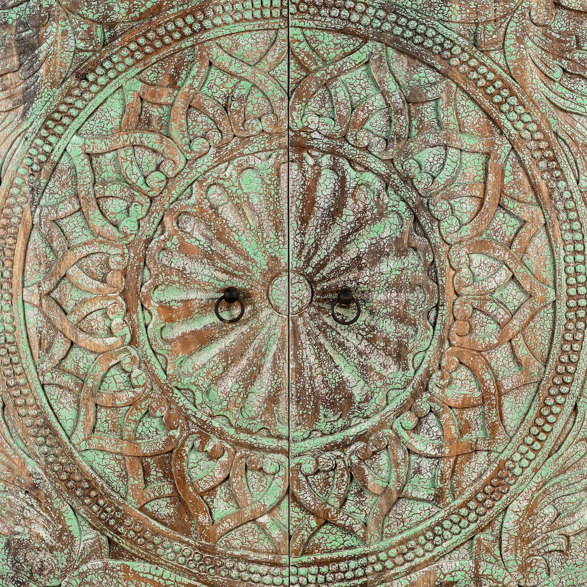Close up image of a carved mango wood cabinet with floral hand-carving, distressed green finish, iron stand, 100 × 40 × 150 cm.
