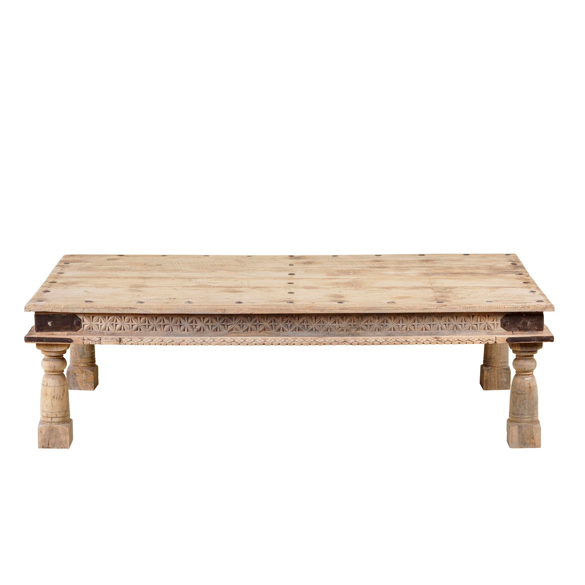White background image of a rustic coffee table Ritvik made from old reclaimed teak wood, featuring a weathered finish, turned legs, decorative carved apron, and visible metal rivets, styled with earthy decor in a minimal interior setting.