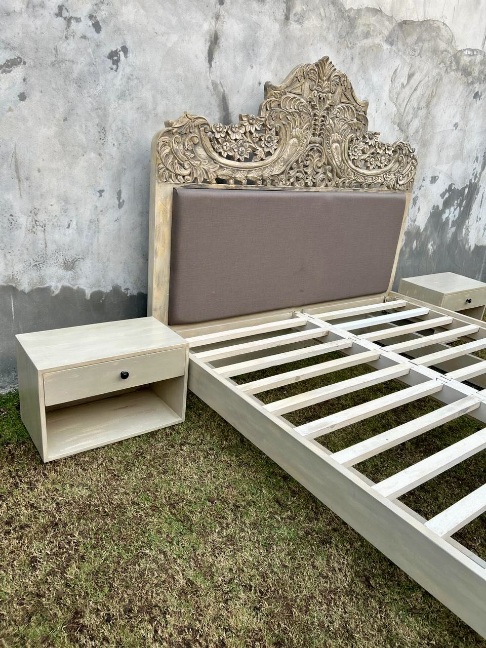 Factory image of a hand-carved vintage bed Samar made from distressed mango wood, featuring an ornate high headboard with soft grey fabric upholstery. A unique blend of Indian craftsmanship and contemporary comfort in a natural, neutral palette.