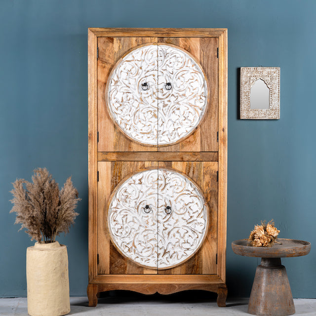A hand-carved mango wood cabinet with floral designs, placed against a blue wall next to a decorative mirror and a vase with dried plants.