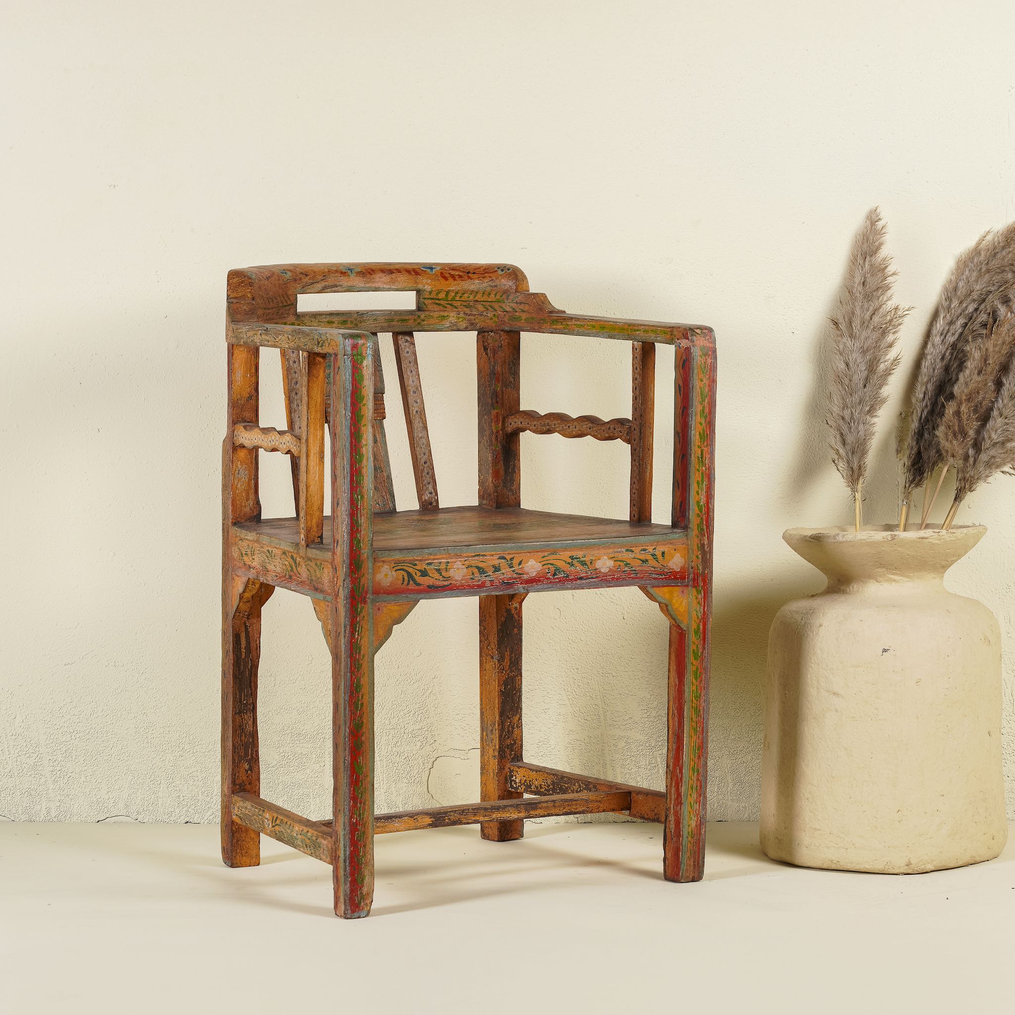 Side angle image of a hand-painted armchair crafted from reclaimed teak wood, featuring intricate traditional patterns in earthy tones on the backrest, seat, and frame, with a rustic and vintage finish.