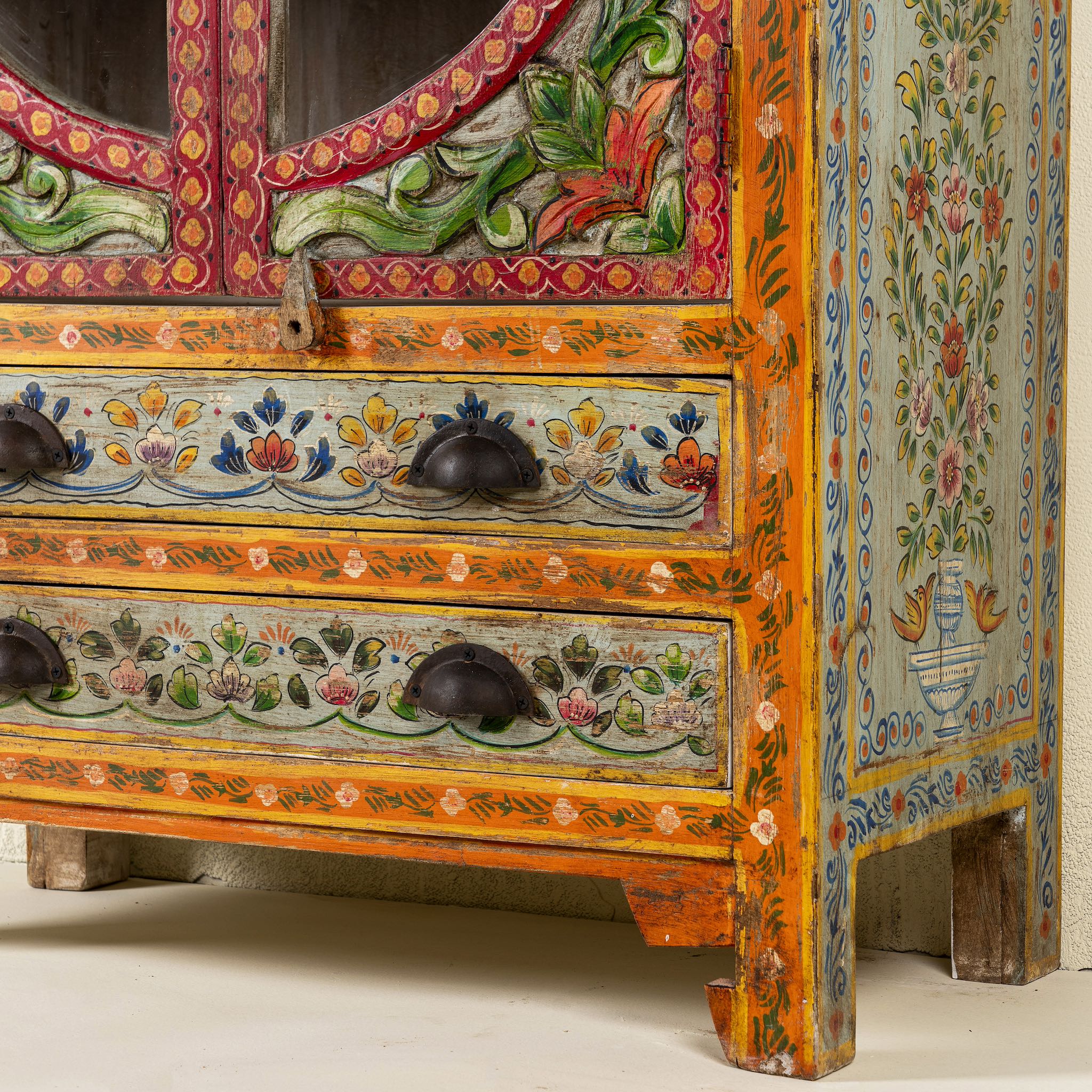 Close up view of the leg part of a hand-painted showcase cabinet made from mango wood, featuring a circular glass-front display, floral motifs in rich orange, green, and red tones, and five drawers for added storage.