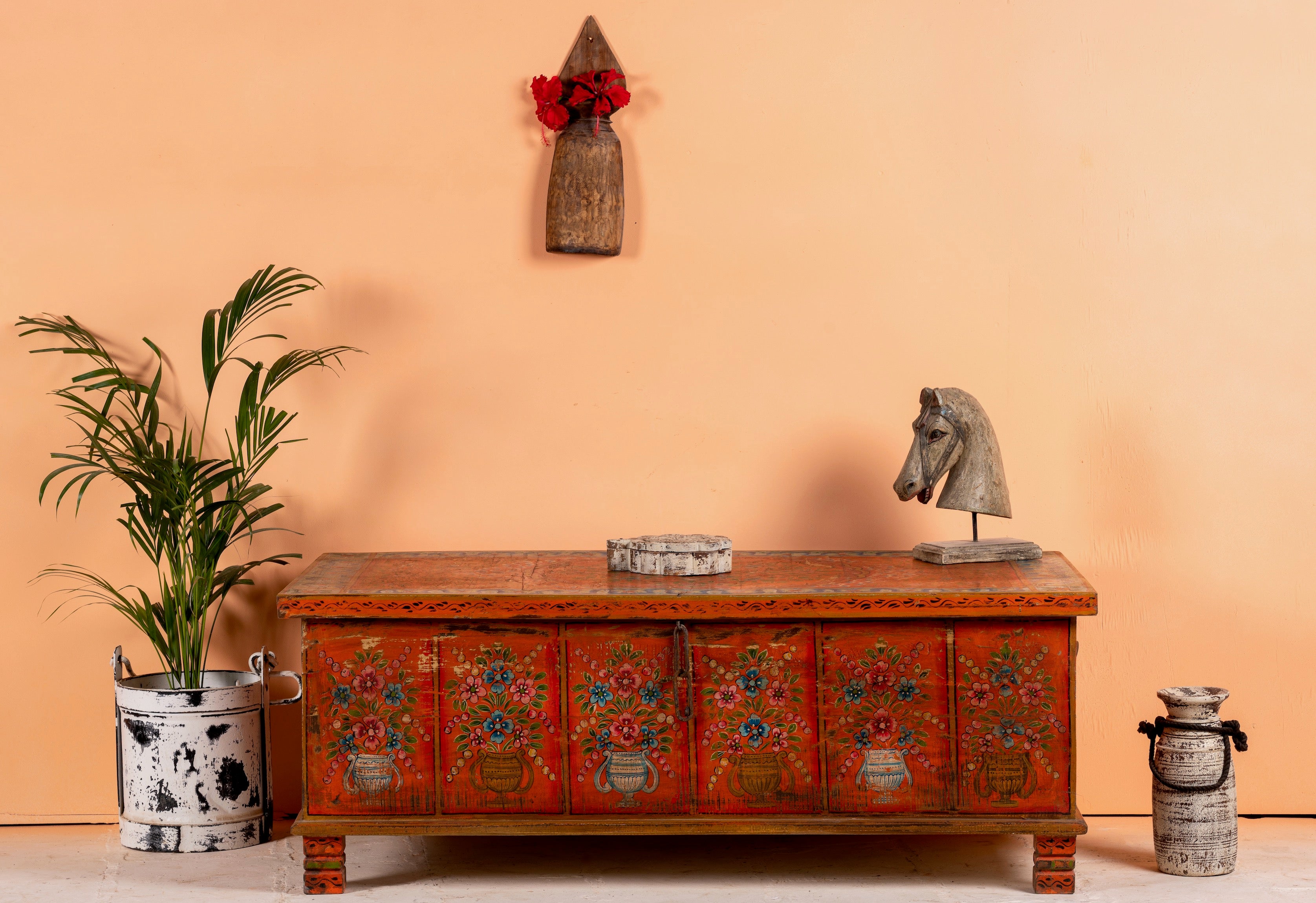 A hand-painted wooden box table with floral patterns, placed against an orange backdrop with decorative items on and around it, including a potted plant and a sculpture.