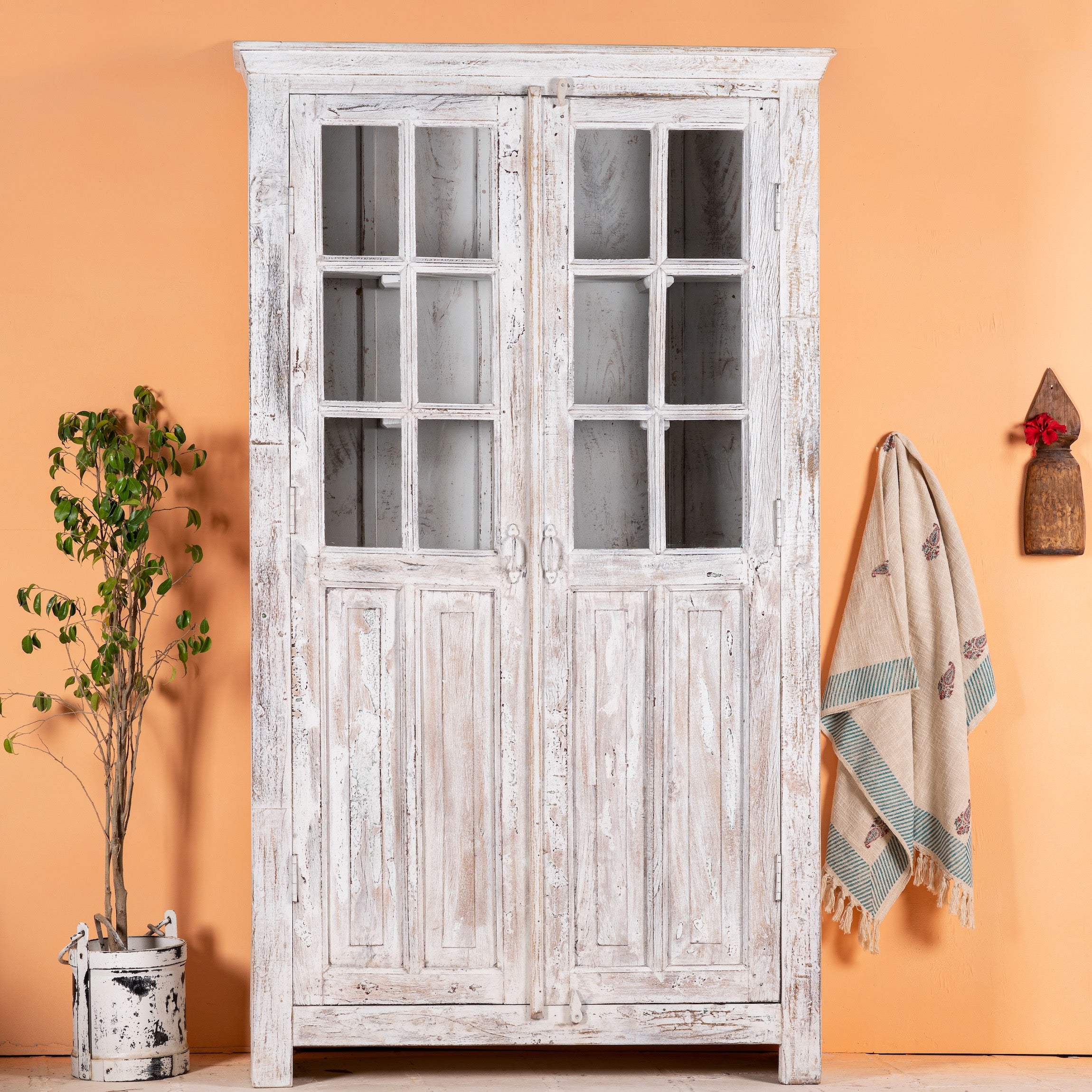 A refurbished antique teak wood showcase with white washed finish, featuring glass panelled doors and a whitewashed interior, standing against a plain wall with a hanging plant and a traditional Indian textile behind it.