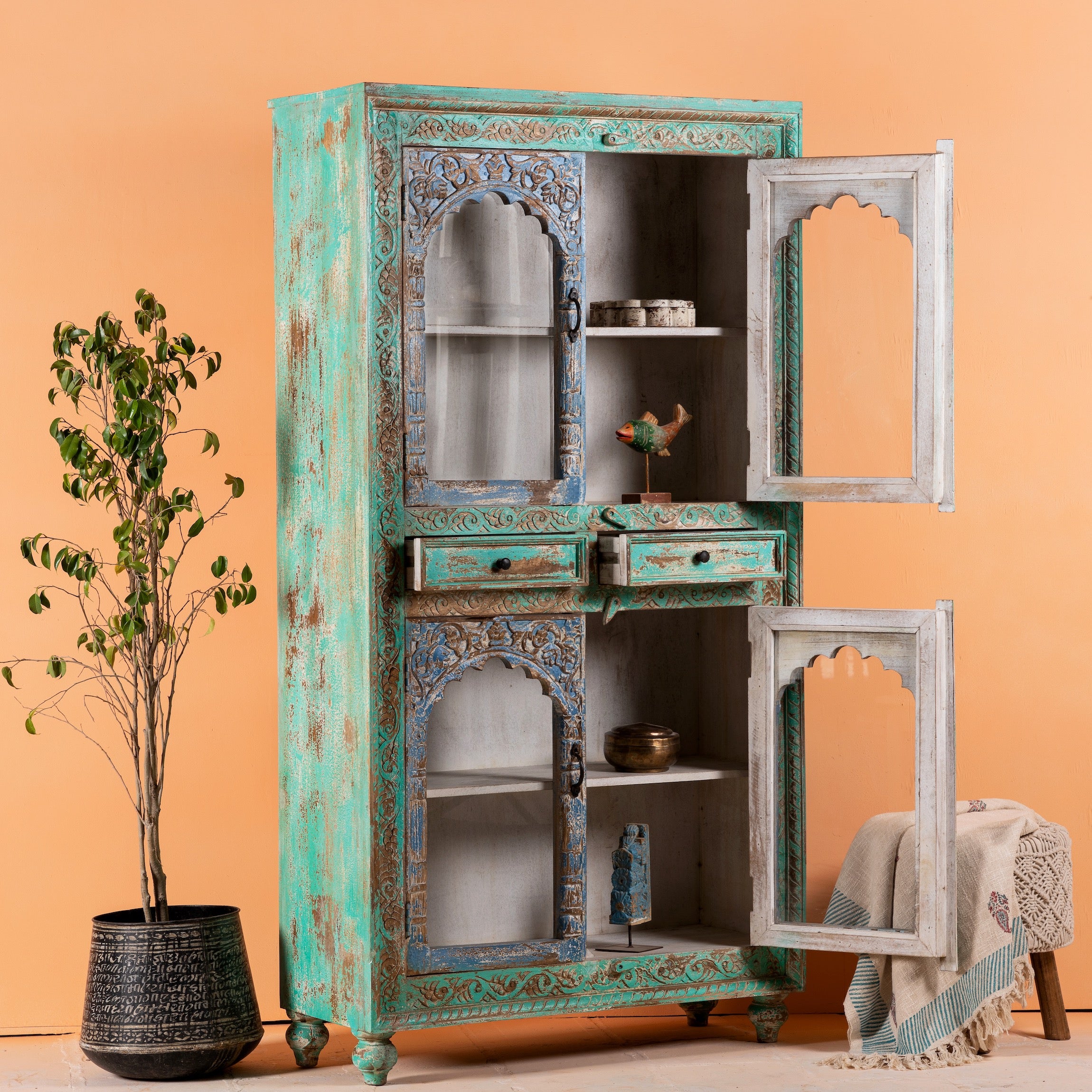 Side angle image with open door of a hand-carved mango wood showcase with glass doors and drawers – carved wooden furniture in distressed turquoise and blue, combining artisan detail with vintage charm.