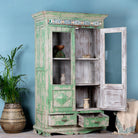 Side angle image with open door of a reclaimed teak wood vitrine with distressed green finish, ceramic inlays, and glass-panelled doors – sustainable furniture with vintage Indian charm.