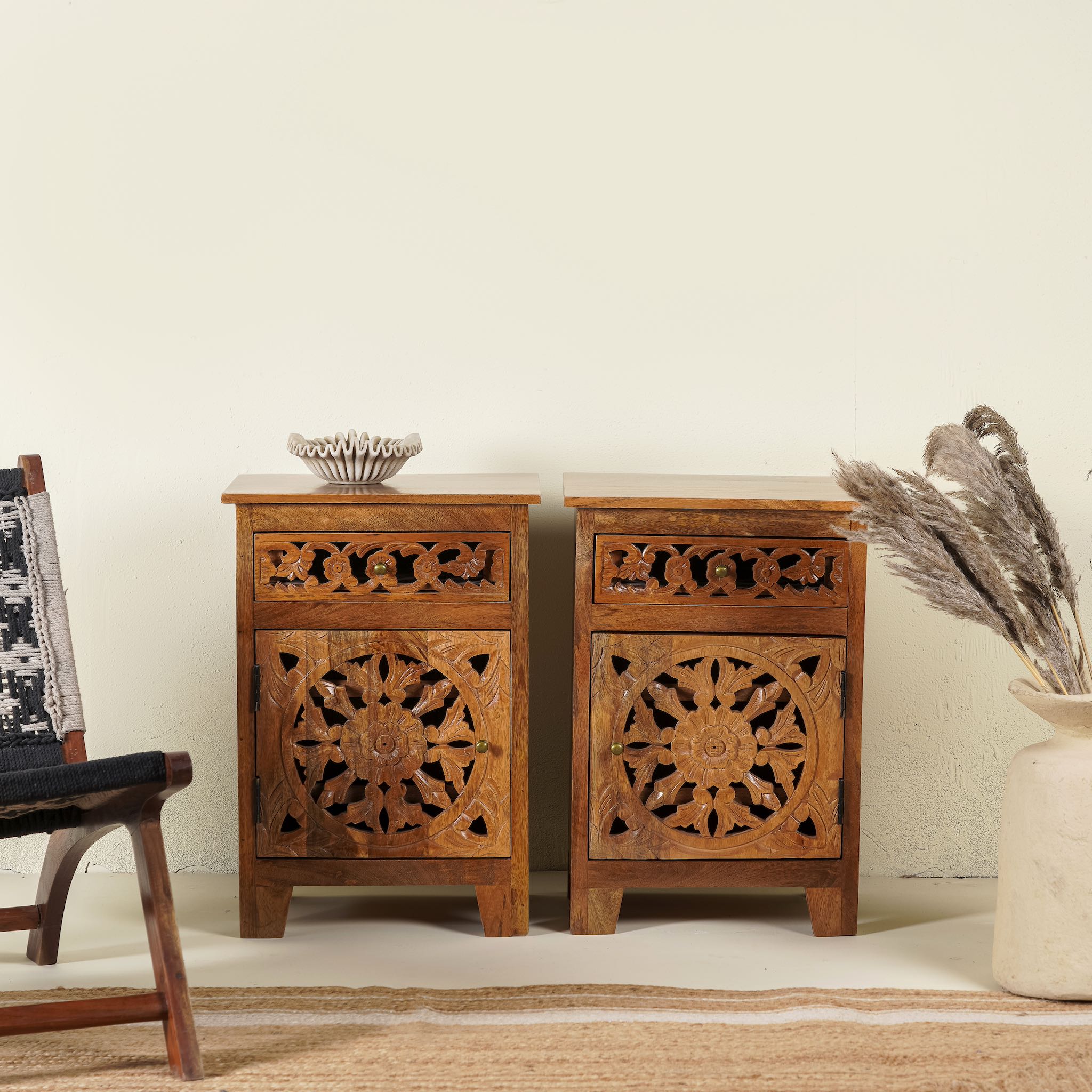 A pair of hand carved mango wood bedside tables with intricate floral and geometric carvings, finished in a warm teak colour, displayed in a room setting with decorative items on top.