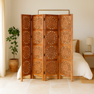 Lifestyle image of a foldable room partition made of mango wood, featuring four panels with symmetrical hand-carved circular floral medallions, finished in a rich natural wood tone and connected by black metal hinges for easy folding and adjustment.
