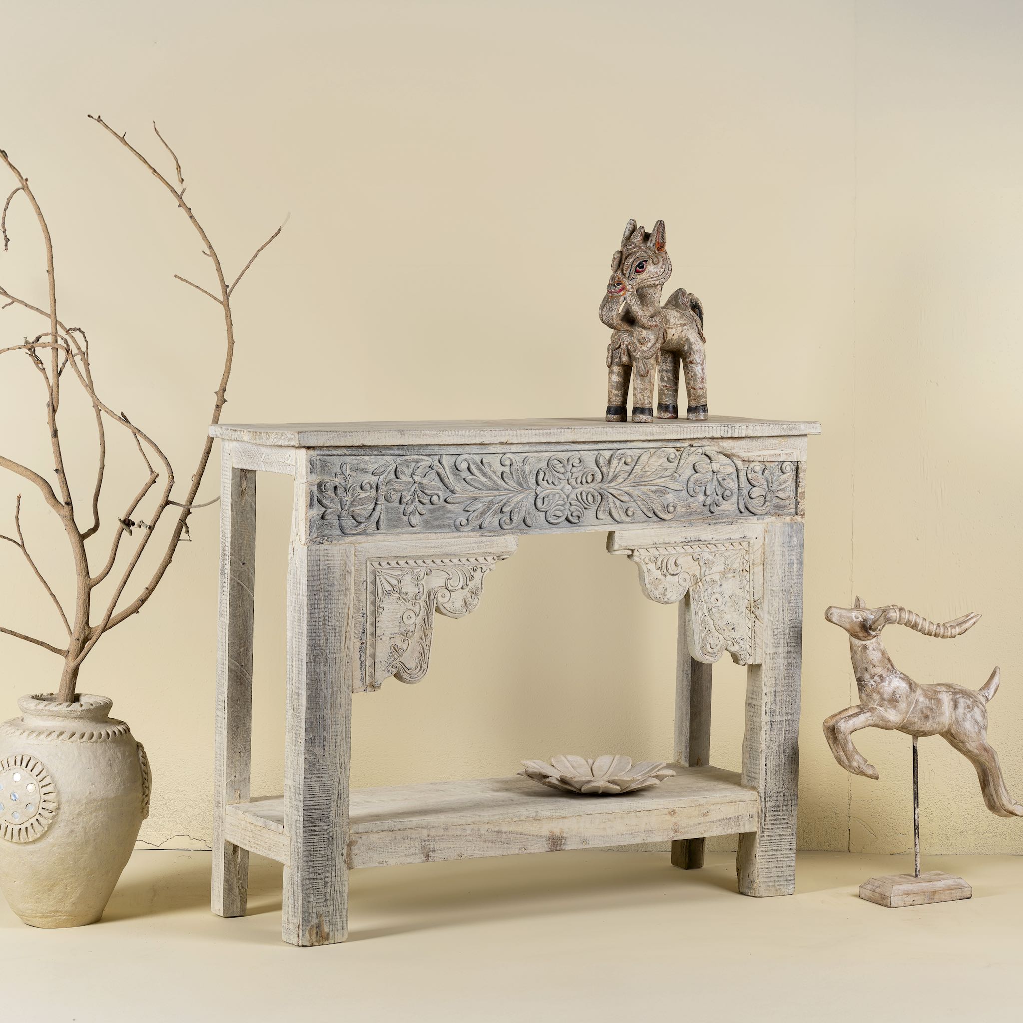 Side view image of a refurbished teak wood console table with a whitewashed finish, featuring intricate hand-carved floral and scroll detailing on the front apron and corners, and a lower shelf for added display or storage.
