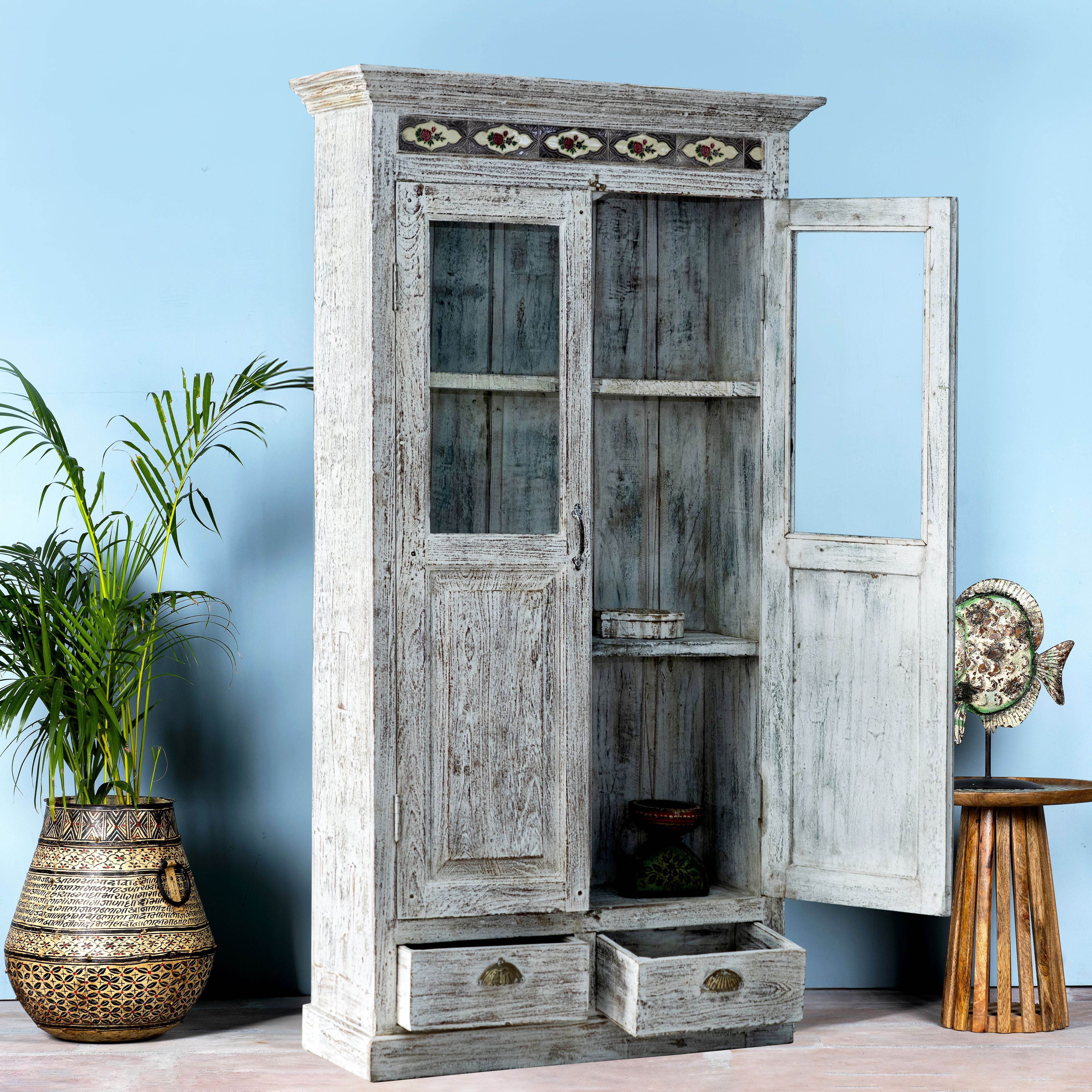 Side angle image with open door of a reclaimed teak wood cabinet in shabby chic white with glass doors and ceramic inlays – dining room furniture with vintage elegance.