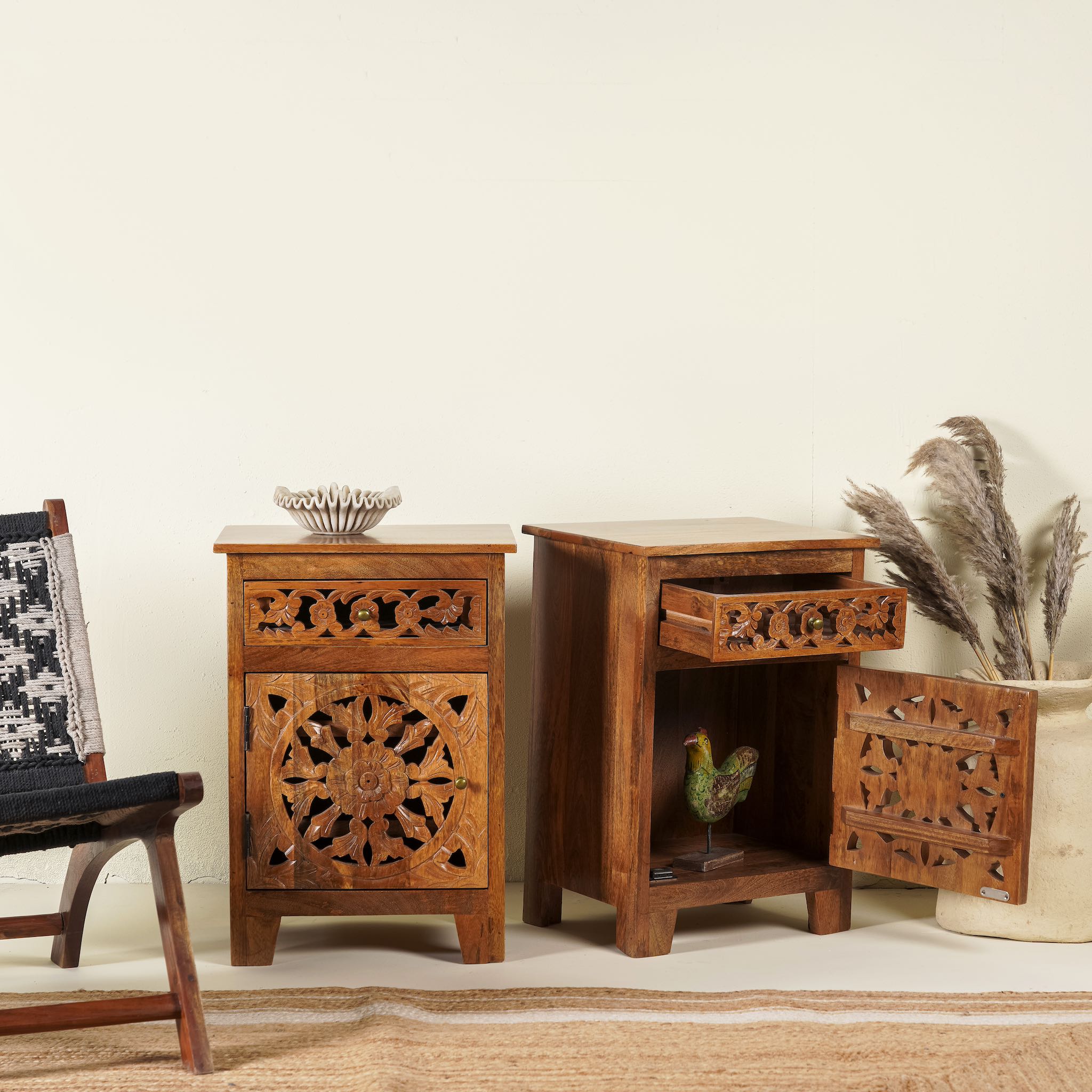 Side angle image of a pair of hand-carved bedside tables made of mango wood, featuring intricate round floral hand carvings on the doors. 