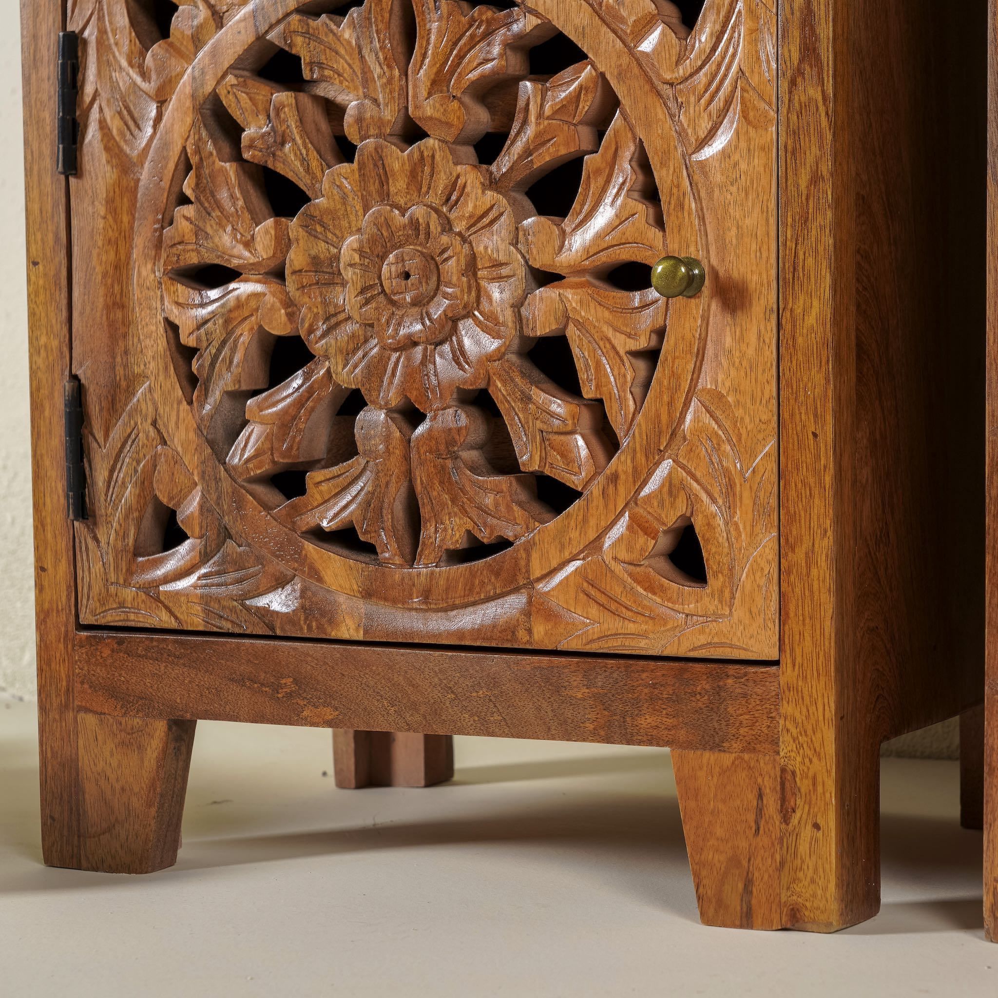 Close up image of the leg part of a pair of hand-carved bedside tables made of mango wood, featuring intricate round floral hand carvings on the doors. 