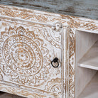 Close up picture of a low board made from solid mango wood, featuring intricate hand-carved mandala motifs, whitewashed finish, and practical storage with two doors and open shelving.