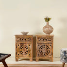Two hand carved mango wood bedside tables with intricate floral and geometric patterns, one with a vase on top and the other with a bowl.