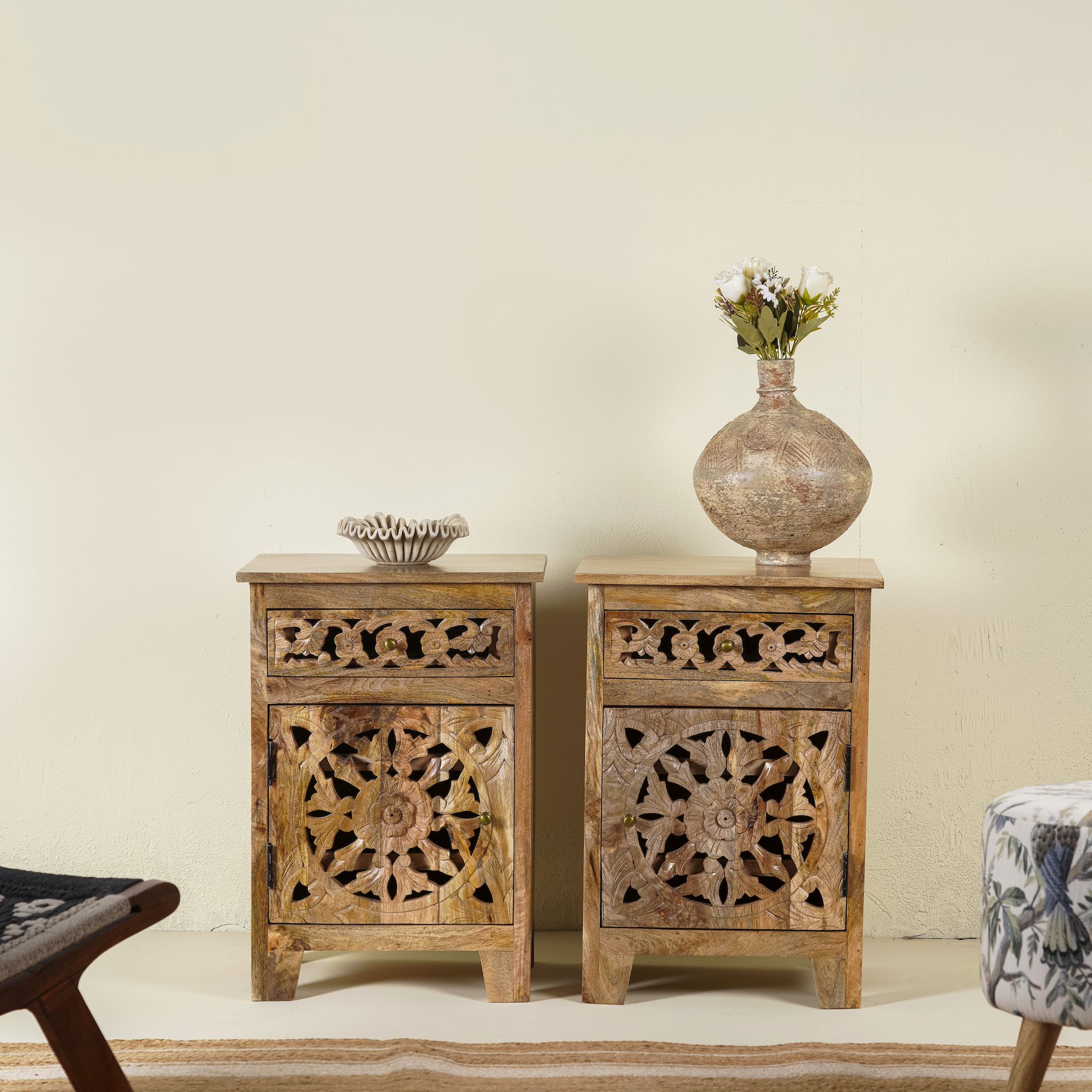 Two hand carved mango wood bedside tables with intricate floral and geometric patterns, one with a vase on top and the other with a bowl.