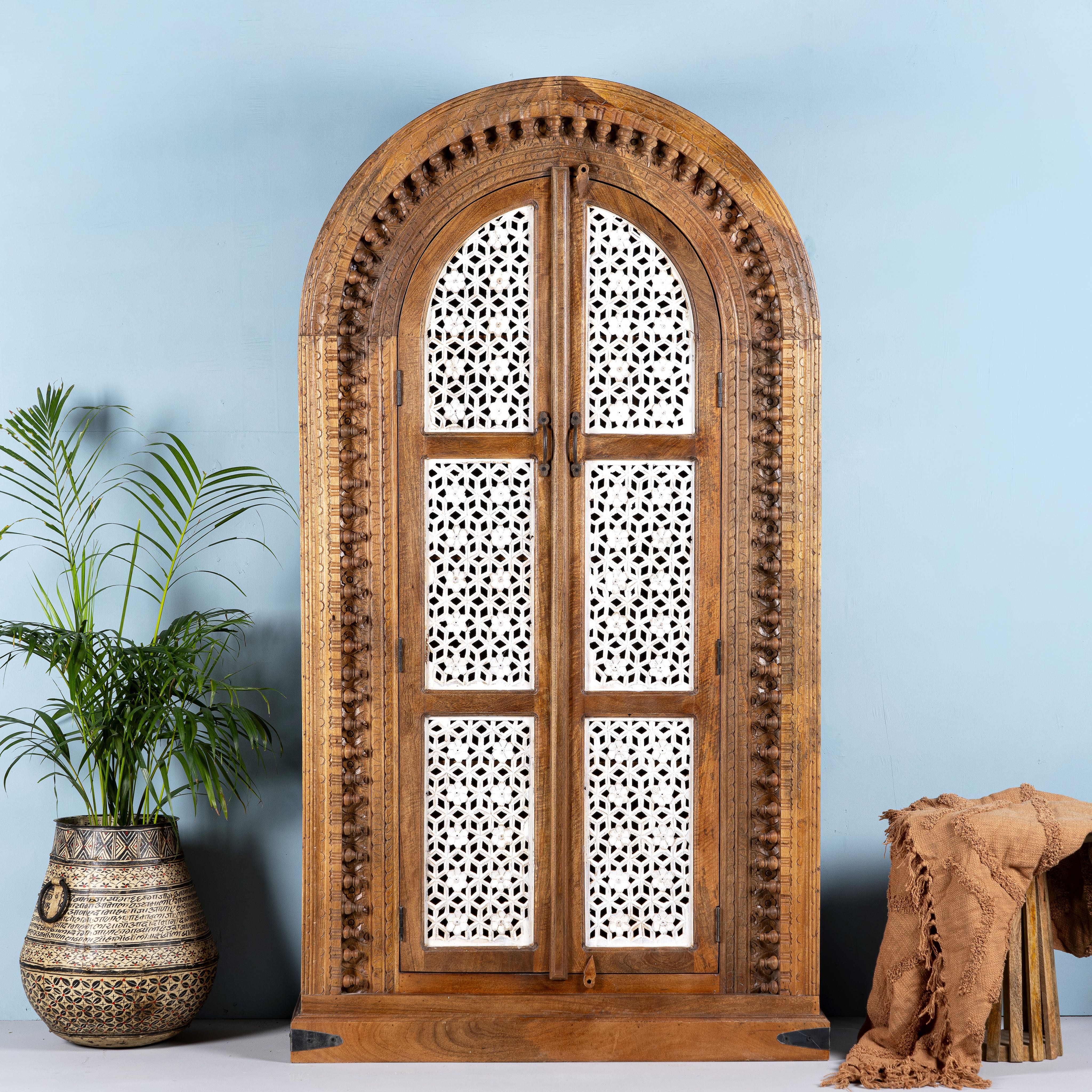 A hand-carved mango wood wardrobe with half-round shape and whitewashed lattice-style panels, featuring intricate carvings. The wardrobe is positioned against a light blue wall, with a potted plant and a brown textured object to the side.
