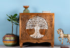 Solid mango wood cupboard with hand-carved Tree of Life design and whitewashed finish, featuring two doors and a curved base, placed against a blue wall with decorative items around it.