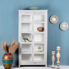 A white, wooden teak wood showcase cabinet with glass doors and various decorative items displayed on its shelves, placed against a blue wall with colorful vases and mirrors.