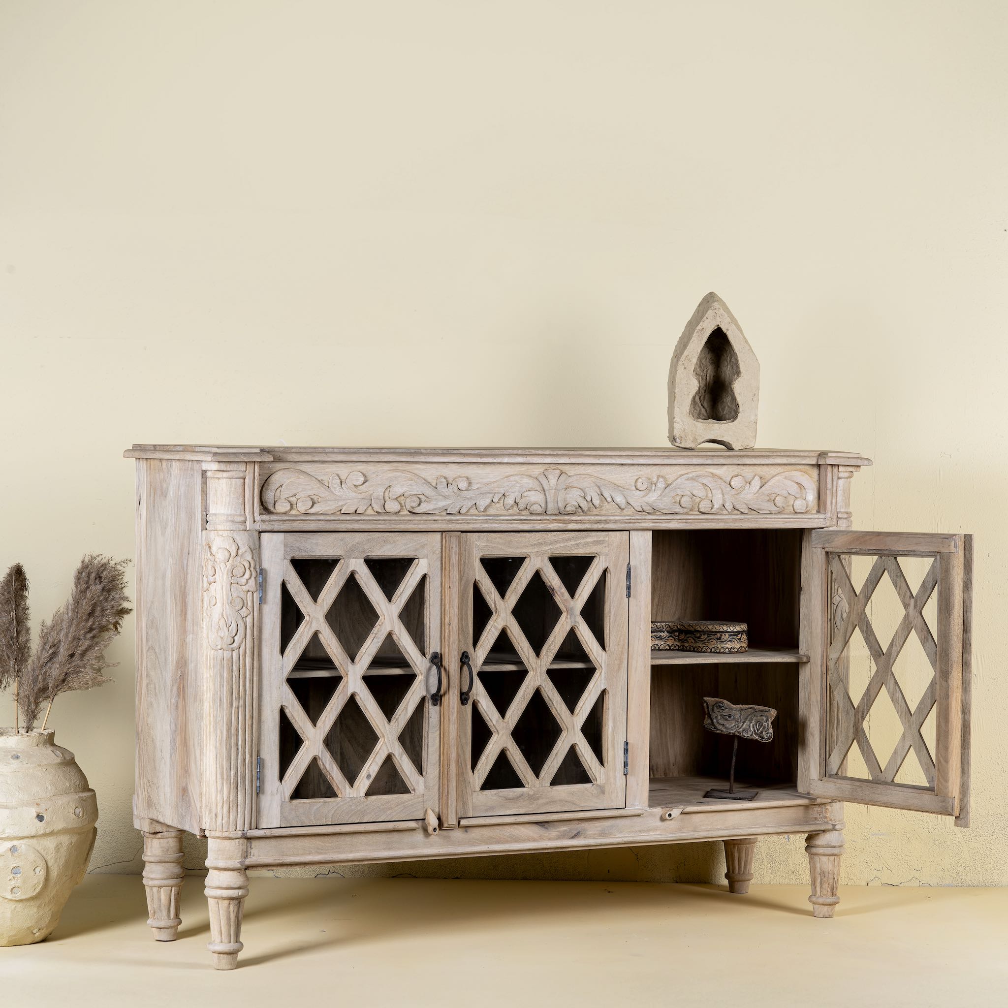 Side angle image with open door of a rustic sideboard made of Mango wood with hand-carved details and glass panelled doors featuring a diamond lattice design.